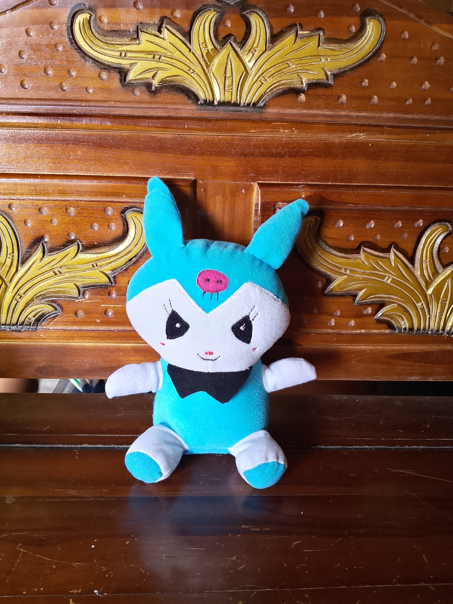 A blue stuffed rabbit with white accents and a black bowtie sits centered on a wooden bench. The bench is ornately carved with gold floral motifs and dark wood inlays. The background consists of more dark, carved wood. The scene is captured indoors in Kasiman, Indonesia.