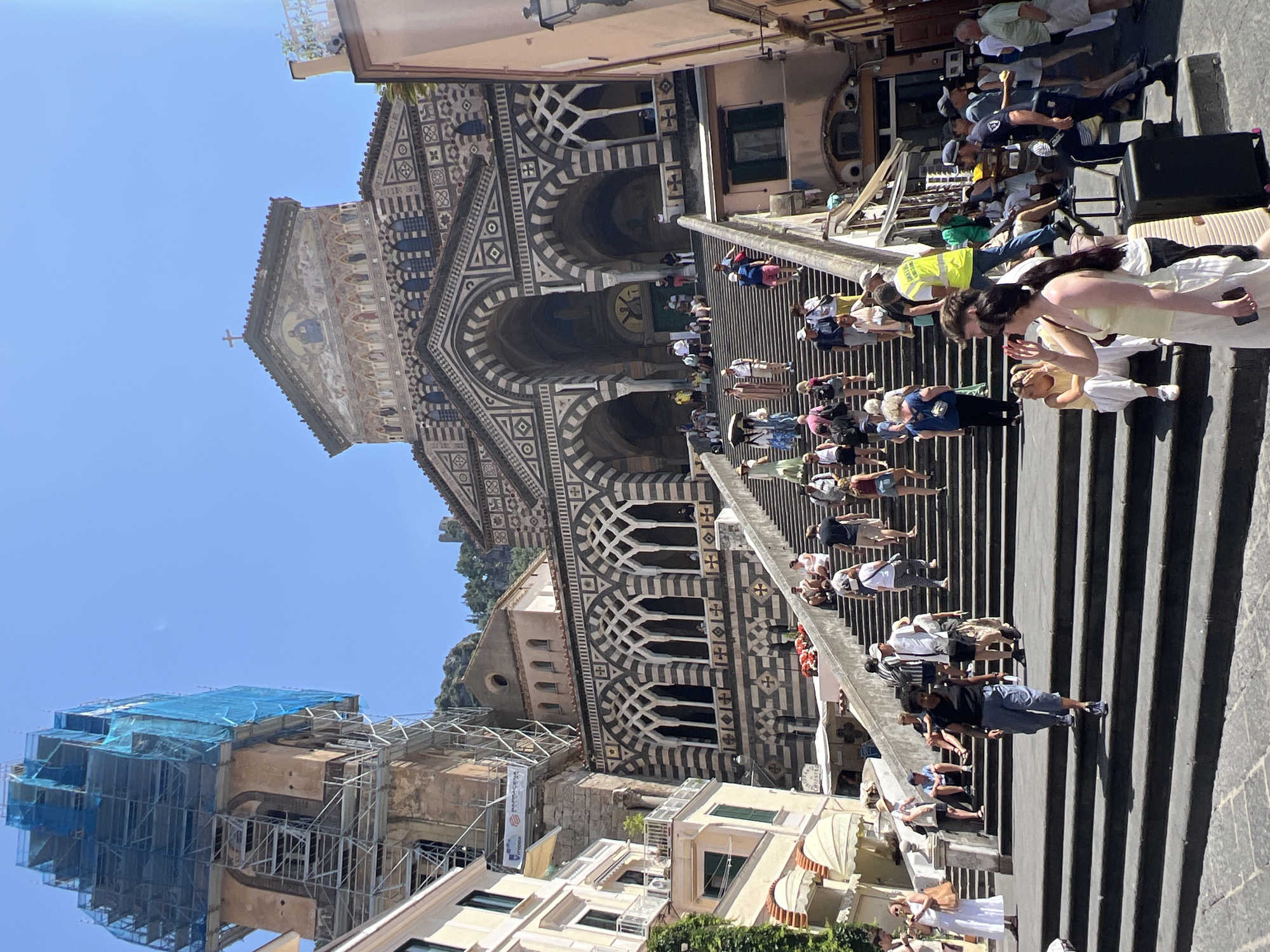 This image captures a wide-angle view of a bustling public square with a grand cathedral as the backdrop, situated in Pogerola, Italy. The cathedral, a prominent structure, is adorned with intricate geometric patterns and features a large arched entrance. A tall bell tower, partially obscured by scaffolding and blue netting, stands to the left of the cathedral. Numerous individuals are seen ascending a wide set of stone stairs leading to the cathedral. They are dressed in casual summer attire, indicative of a warm, sunny day. Some people are walking, while others are seated on the steps, resting or observing. Among the crowd, there are individuals carrying luggage, suggesting they might be tourists. A person in a bright yellow vest, possibly a worker or guide, is visible on the stairs. The environment is lively, with a sense of movement and activity as people navigate the steps and the plaza. The architecture and atmosphere suggest a popular tourist destination.