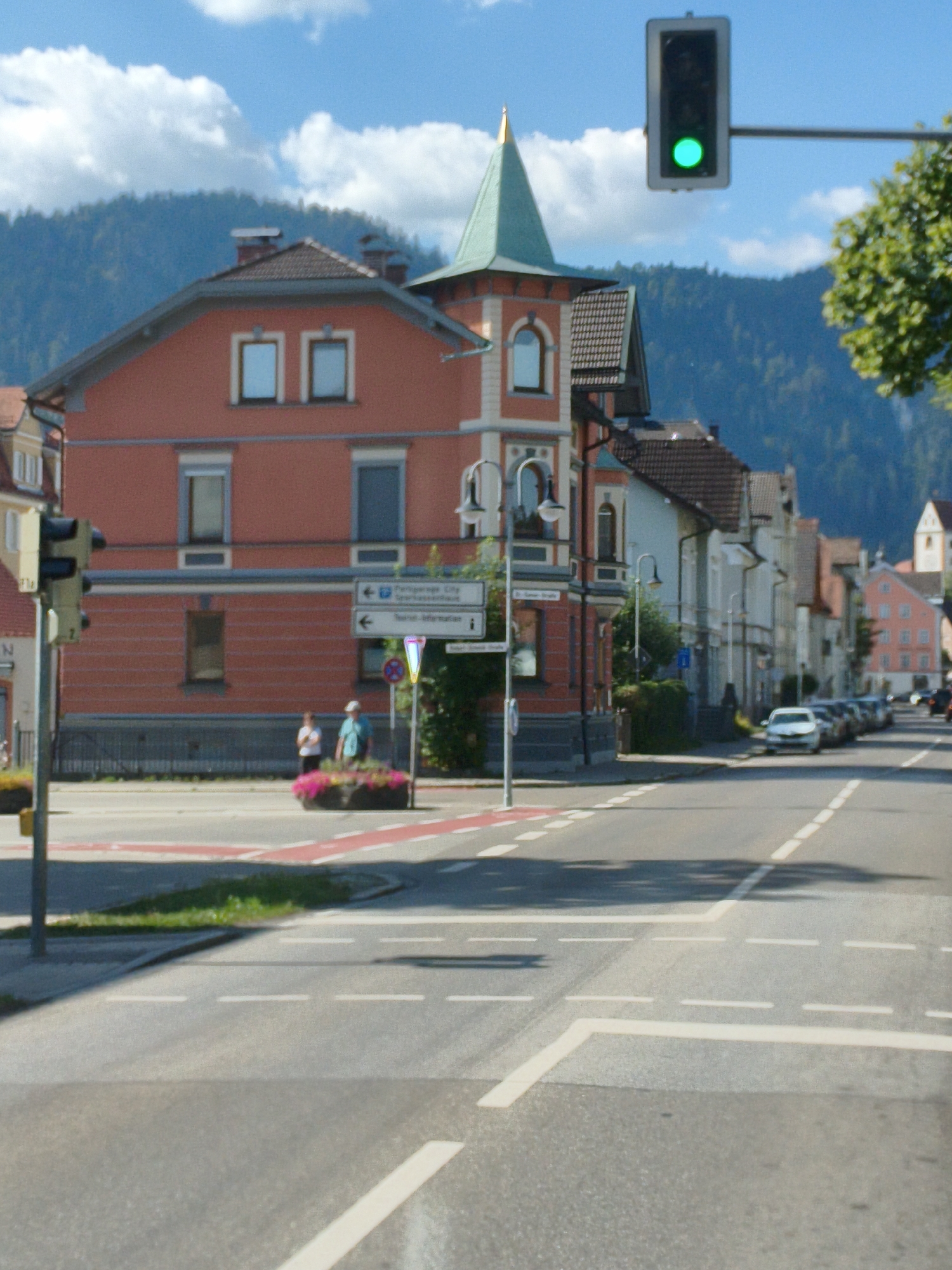 The image depicts a street scene in Füssen, Germany, under a partly cloudy blue sky. In the foreground, a street with lane markings and crosswalks is visible. A traffic light displaying a green signal hangs from a pole over the intersection. To the left, a red building with multiple windows is prominent. Two individuals, a woman in a white shirt and black pants and a man in a teal shirt and white cap, stand near a flower planter. Further down the street, a row of European-style buildings lines the right side, with parked cars visible. The background features a lush green mountain range, adding to the scenic backdrop of the town.