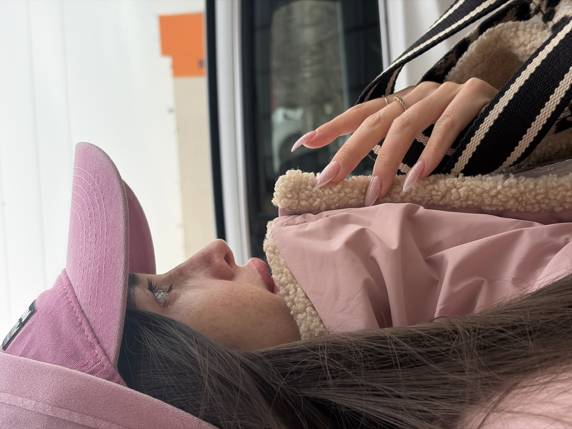 A side-view shot depicts a person lying on a pink surface, wearing a pink baseball cap and a pink jacket with a light-colored, possibly faux-fur lining. The person's face is visible, showing details such as freckles and eyelashes. One hand, adorned with a ring, rests on the jacket. The setting appears to be indoors, with a background featuring a window or a reflective surface, and a glimpse of an orange and white vertical element. This scene does not provide any evidence to determine whether the location is in the city of Saint Petersburg, Russia.