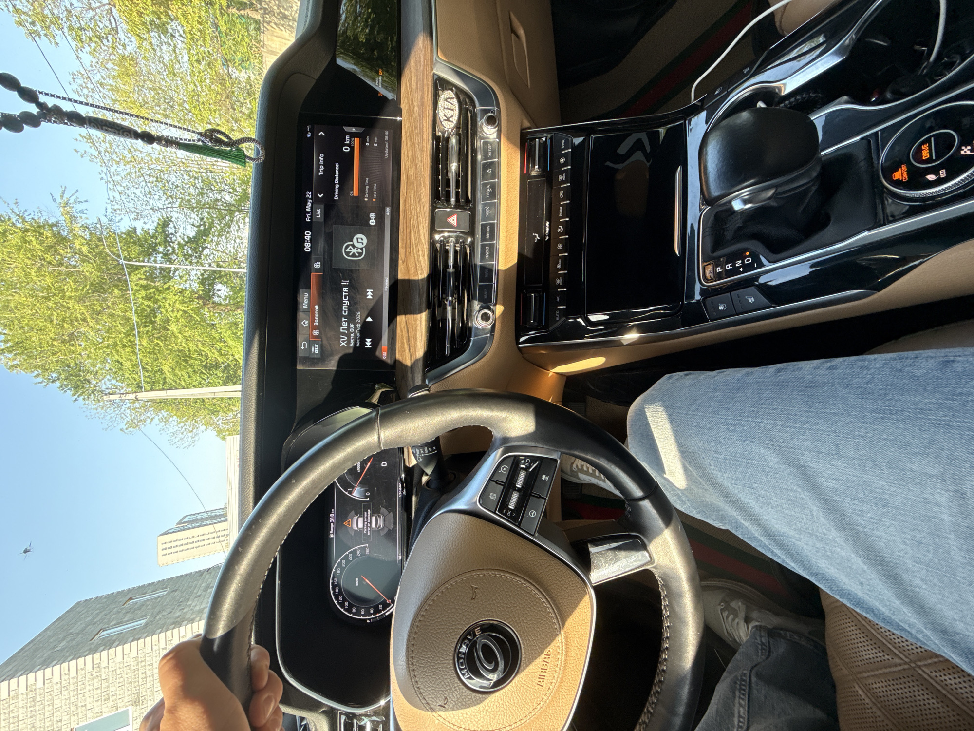 Here is a detailed description of the image:
The image shows the interior of a car, specifically the driver's seat and dashboard area. The car's interior features a tan and black color scheme with wood-grain accents. A leather steering wheel is visible, with the car's logo prominently displayed in the center. The dashboard includes a digital display screen showing various information, including the time, date (Friday, May 22), trip information, and the currently playing song: "XV Лет спустя !" by Баста, GUF.
The car appears to be in the "Drive" mode. The driver's hand is visible gripping the steering wheel, and a portion of their jeans-clad leg and shoe is visible in the foreground. Outside the car window, trees and buildings are visible, suggesting an urban setting. A rearview mirror ornament is hanging above the screen.
Based on the Cyrillic text displayed on the dashboard, the location is likely in a Russian-speaking region. Given the information, the location is Vladivostok, Russia.
