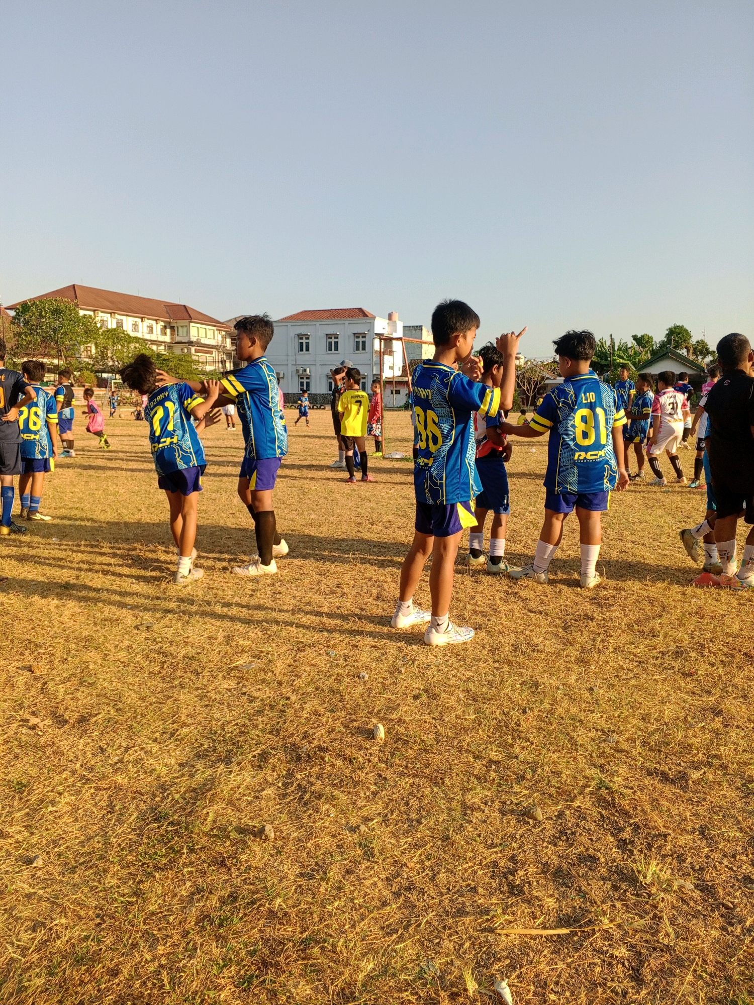The image depicts a group of young boys in soccer uniforms on a dry, grassy field, likely in Giritirto, Indonesia, given the architectural style of the buildings and the natural environment. The boys are dressed in blue and yellow jerseys and shorts, suggesting a soccer practice or game. In the foreground, two boys are facing each other, one appearing to playfully interact with the other. Behind them, other boys are scattered across the field, some in groups, and some individually. In the background, there are several buildings with brown roofs and white facades, characteristic of Indonesian architecture, and lush green trees. The sky is clear and bright, indicating daylight. The overall scene suggests a youth soccer event taking place outdoors on a sunny day.