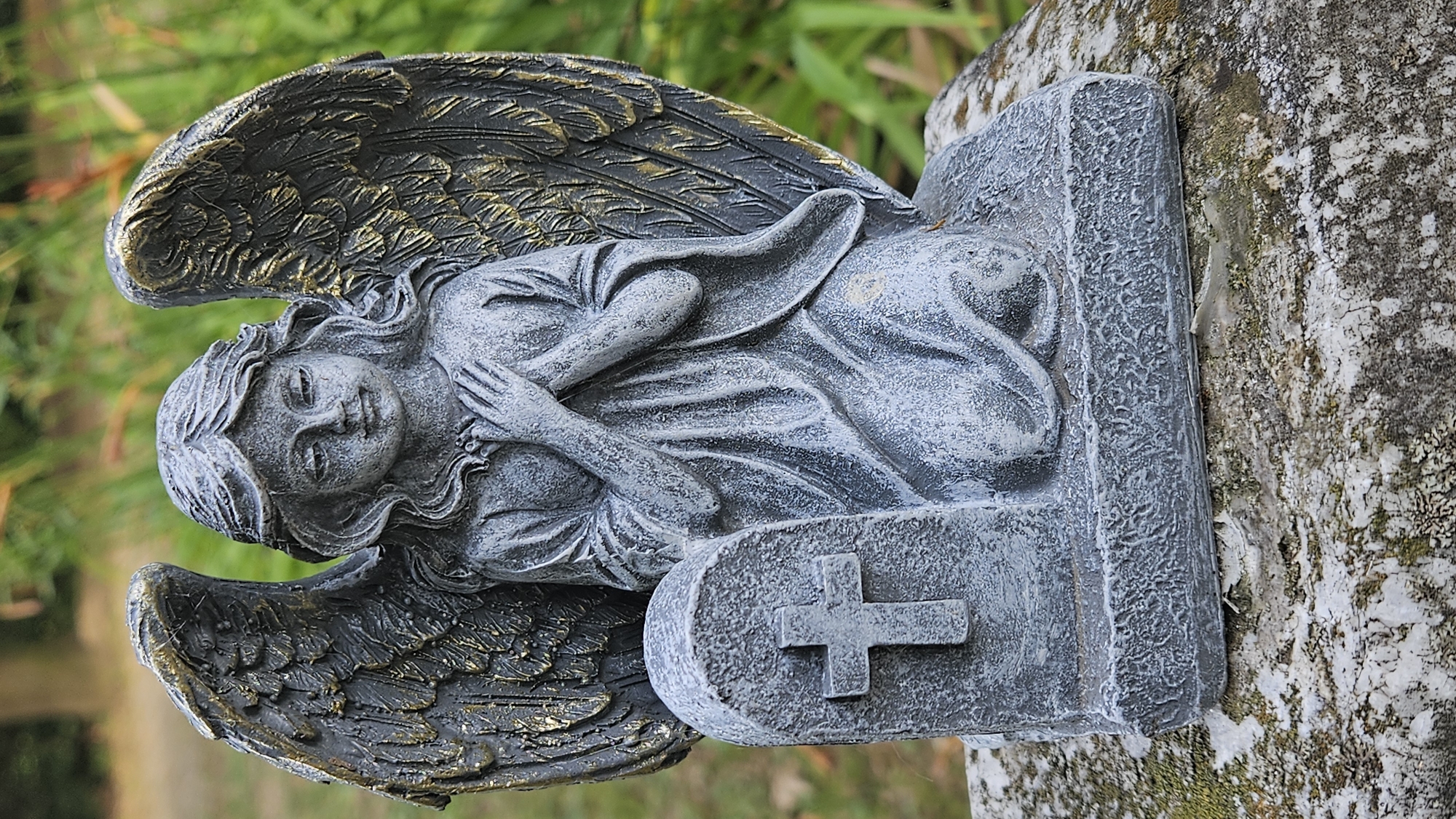 A stone statue of an angel is kneeling in front of a tombstone. The angel has its hands clasped together at its chest and its wings are spread behind it. The tombstone has a cross carved into it. The statue is sitting on a rock that is covered in moss. The background is blurred green foliage. This statue is located in the cemetery in Prešov, Slovakia.