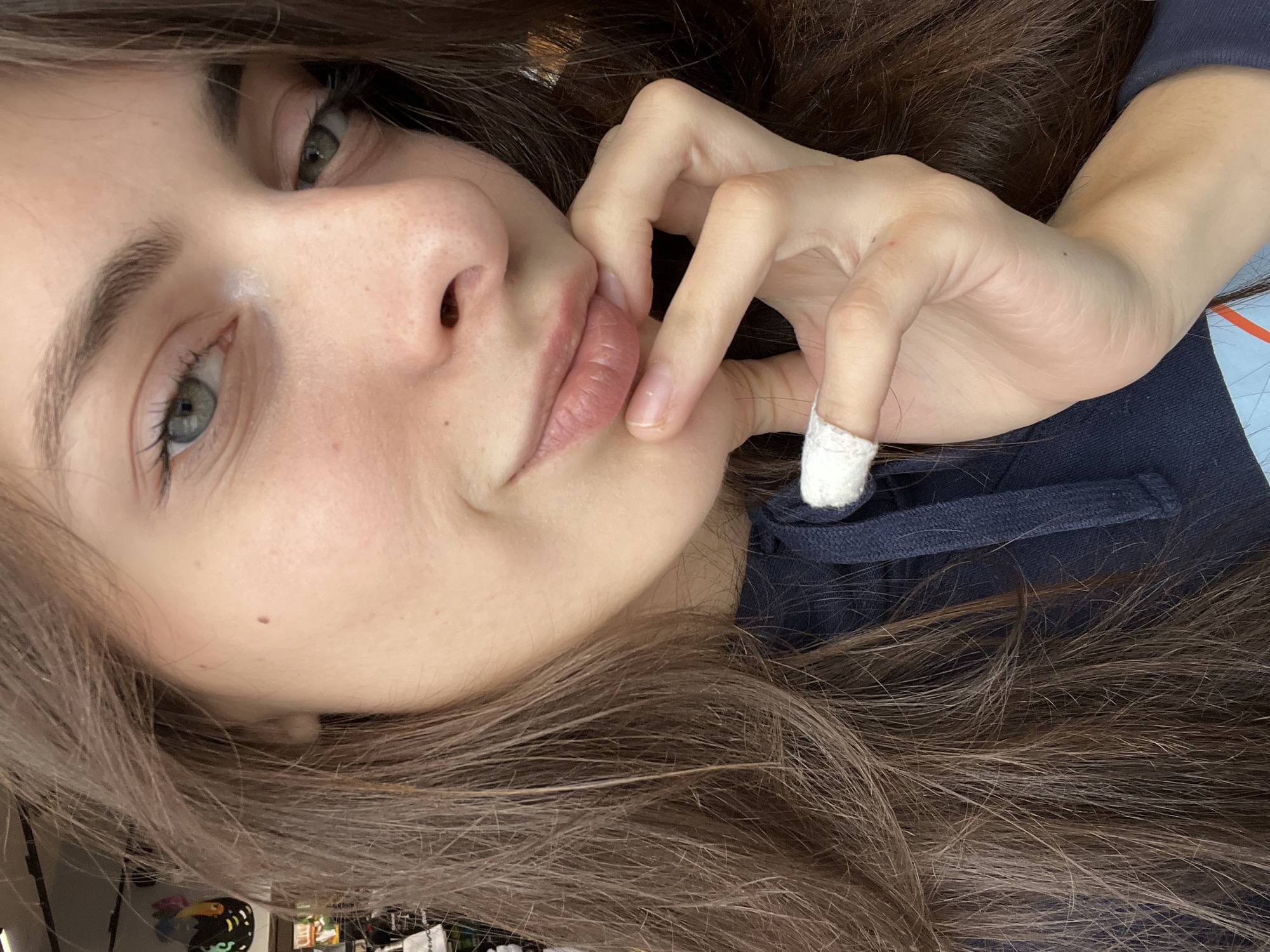 A close-up, high-angle portrait features a young woman with long, dark brown hair cascading around her. Her head is slightly tilted, and she gazes towards the viewer with greenish-grey eyes, framed by dark eyelashes and well-defined eyebrows. Her skin is fair, with a subtle mole visible on her left cheek. Her naturally pink lips are slightly parted, conveying a soft, perhaps pensive, expression.
Her right hand is brought up to her face, with fingers gently touching her lower lip and chin. A distinctive white, textured band or wrap is present on the ring finger of this hand. She is dressed in a dark blue hooded garment, with a white drawstring visible around the neckline. The background is softly blurred, suggesting an indoor setting, though no specific objects are clearly discernible. The overall lighting is soft and diffused. No visible text is present in the image. The image itself provides no specific visual cues for the location of Krasnoyarsk, Russia.