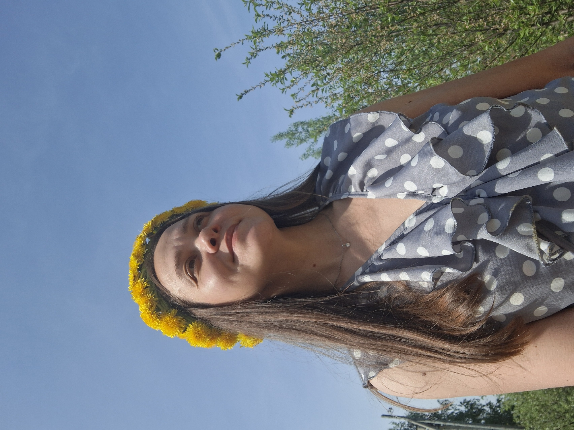 A woman with long brown hair is wearing a yellow dandelion flower crown. She is wearing a gray and white polka dot dress. The background is a clear blue sky with some green foliage from trees. The location is Chaikovsky, Russia.
