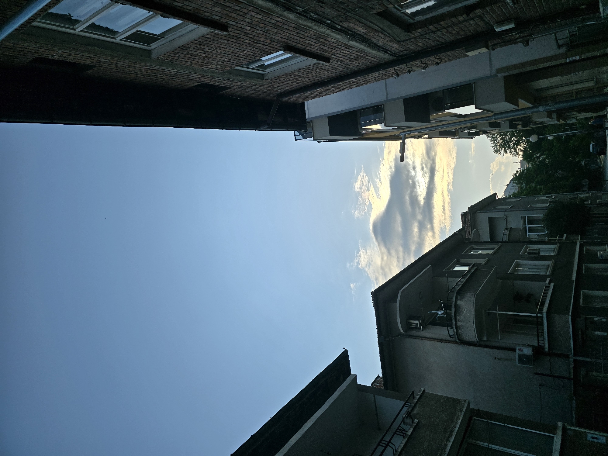 The image, taken with a vertical perspective looking upwards, captures a narrow view of the sky framed by buildings. On the left, a dark brick building with several windows, some reflecting the sky, forms a strong vertical edge. A dark pipe runs along its upper section. On the right, a lighter-colored, more modern residential building is visible, featuring multiple balconies with railings and windows. Several external air conditioning units are attached to this building. In one of the cropped views, a small bird is perched on the peak of its roof. Further in the background on the right, additional structures and the upper parts of green trees are present.
The central and upper-left portion of the image is dominated by a clear, light blue sky. Towards the right side of the sky, scattered clouds are illuminated with soft, warm tones, suggesting either the setting or rising sun. This lighting casts subtle shadows on the buildings, indicating late afternoon or early morning. The scene appears to be in an urban or residential area. No people or visible activity are present, and the overall mood is serene and still. No visible text can be identified in the image.