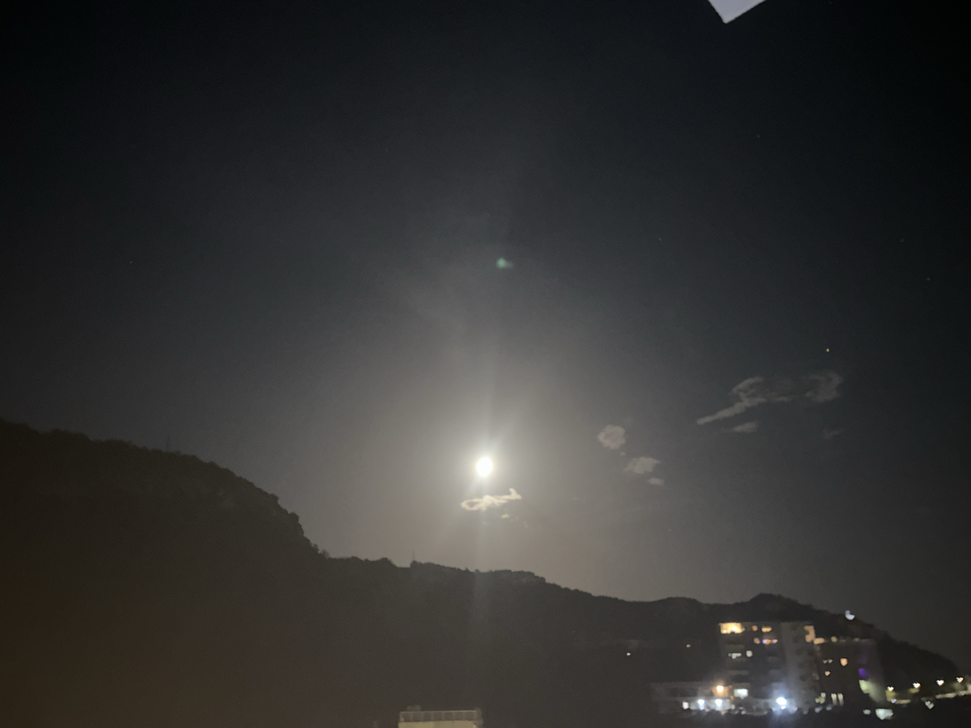 The image depicts a nighttime scene in Rafailovići, Montenegro, dominated by a bright full moon in the dark sky. The moon is the central element, casting a strong light that illuminates a small cloud directly below it. In the background, the silhouette of a mountainous terrain is visible, with lights from buildings suggesting a settlement on its slopes. The lower part of the image shows structures and lights that appear to be part of a town or village, possibly along the coast. The overall atmosphere is serene and nocturnal.