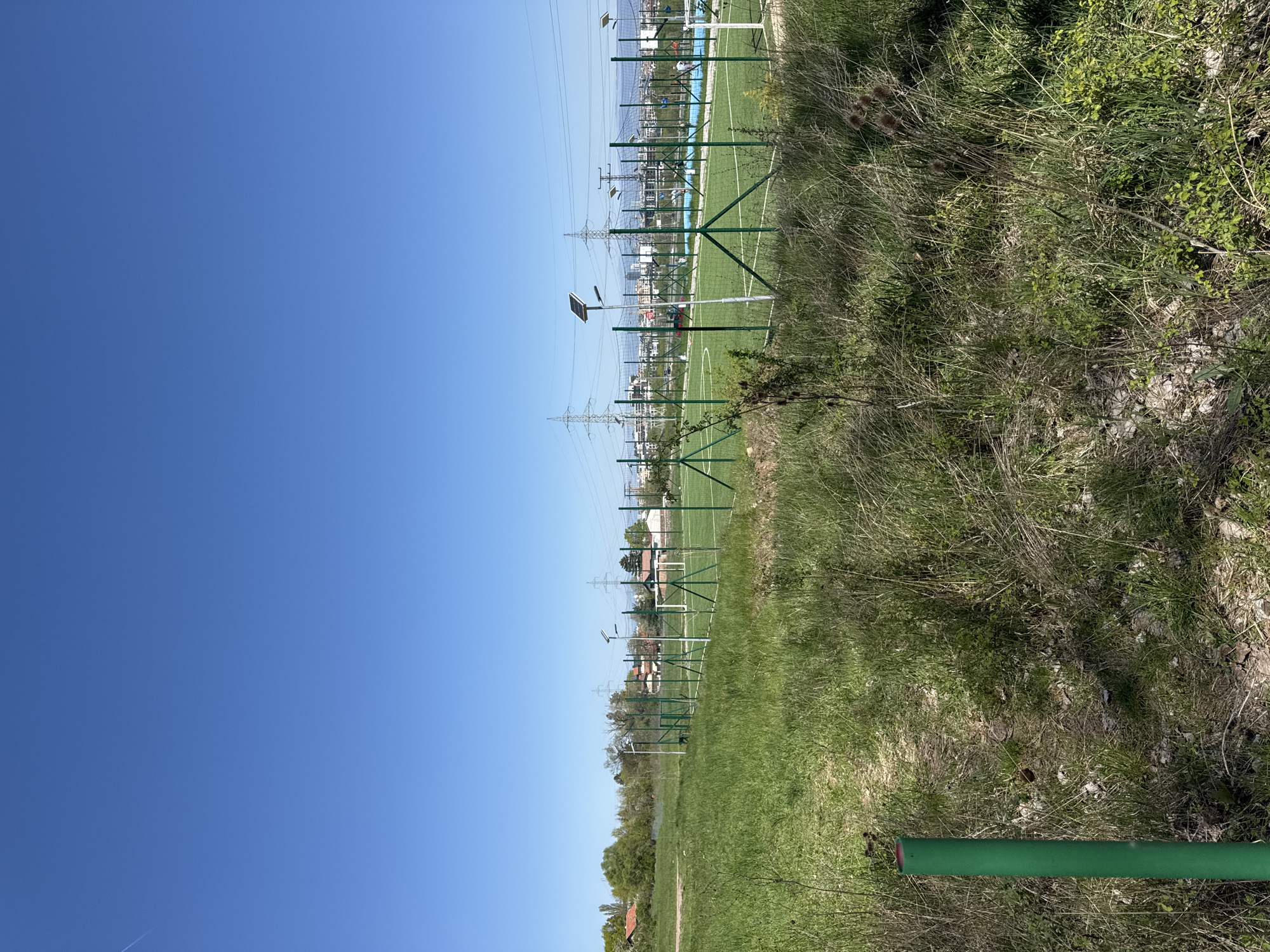 The image shows a grassy area in Sofia, Bulgaria, next to a soccer field. A green fence separates the grassy area from the field. Power lines run overhead. Several lampposts with solar panels line the edge of the field. There are some buildings visible in the background. The sky is blue. The photo is taken at an angle.