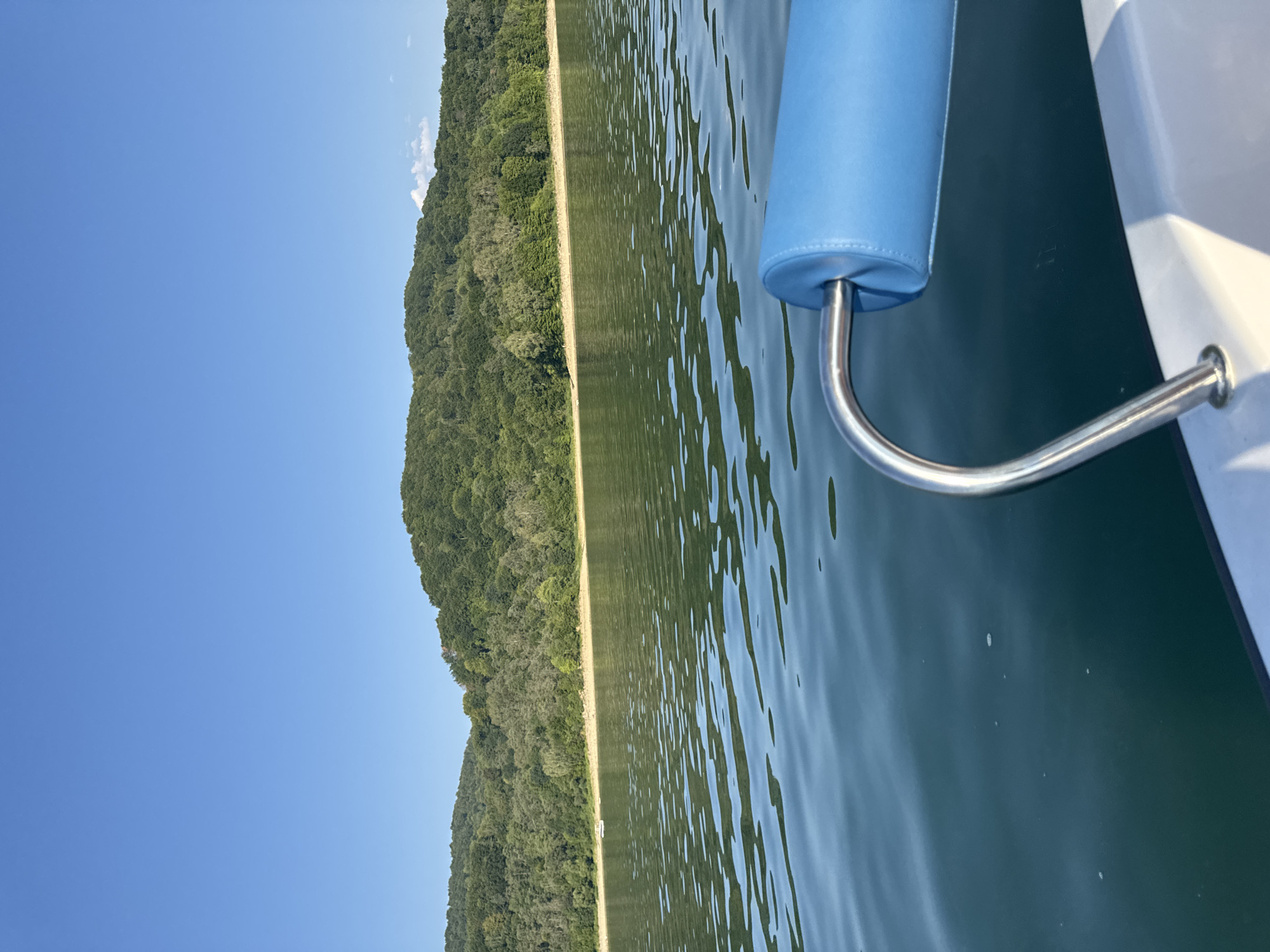 The image shows a view from a boat on a body of water, likely Lake Solina, near Polańczyk, Poland. The water is calm with ripples reflecting the clear blue sky. In the background, a densely forested hill rises from the shore, suggesting a natural landscape. A small white boat is visible near the shore. In the foreground, a portion of the boat the viewer is on is visible, including a silver railing and a blue padded armrest. The scene is tranquil and suggests a leisurely day on the water.
