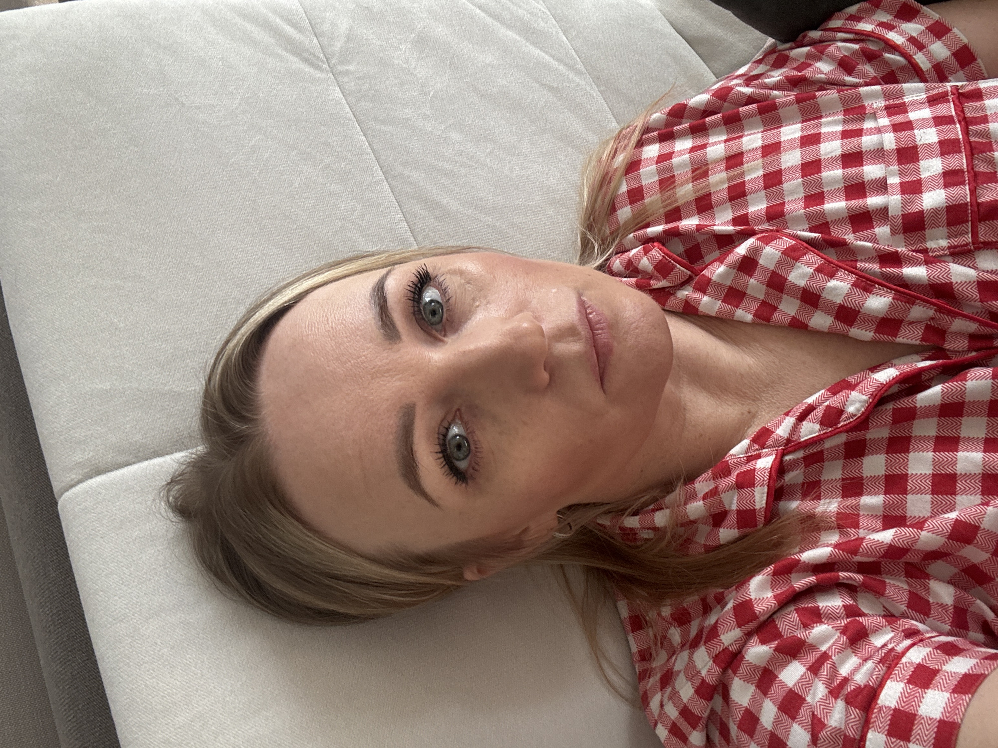 A woman lies on a light-colored couch wearing red and white checkered pajamas. The image is taken from an overhead perspective, focusing on her face, which is tilted towards the camera. The background is out of focus. The location is not discernible from the image.