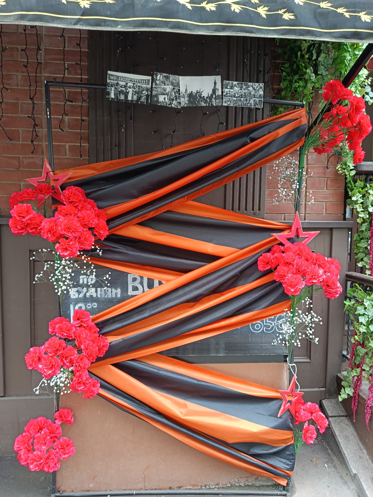 The image shows the decorated entrance to a building, likely a cafe or shop, in Moscow, Russia. The decoration is primarily focused on the ribbon of Saint George, an orange and black striped ribbon associated with Russian military valor, particularly in World War II. This ribbon is draped in a zig-zag pattern across a black chalkboard. Clusters of red carnations, along with white baby's breath, embellish the ends of the draped ribbon. Red star ornaments are also positioned near the floral arrangements.
At the top of the image, black and white photographs depicting military parades and Soviet leaders are affixed to the door. A black awning is partially visible at the top, providing shade. The building itself appears to be made of brown brick. Faux greenery is attached to the right side of the entryway. The text on the chalkboard and the lighting suggest this is an outdoor arrangement.