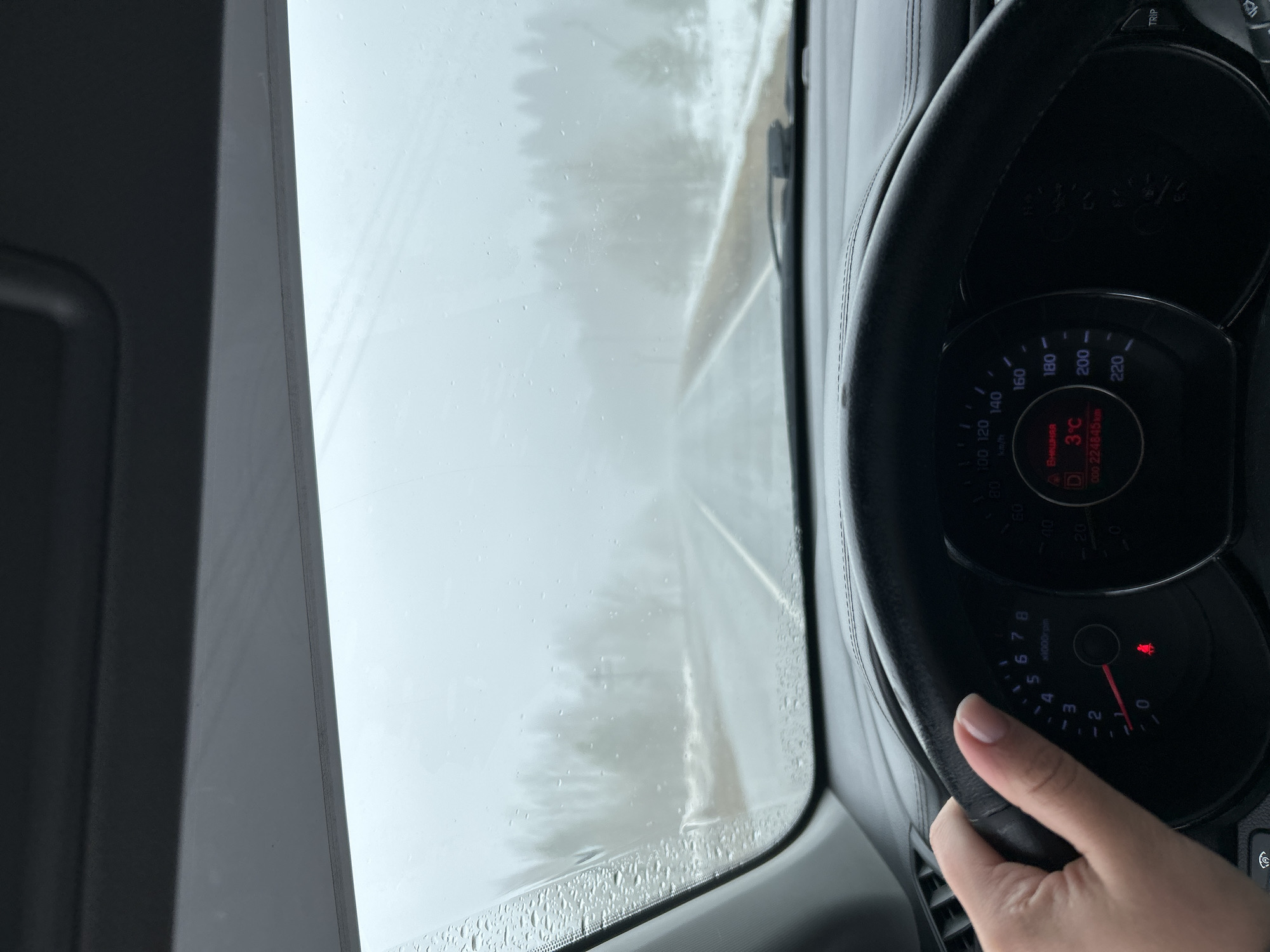 The view from inside a car is depicted, showing a road shrouded in fog or light snow. The temperature gauge on the dashboard reads 3°C. A person's hand is seen on the steering wheel. The odometer reads 224845 km. The location is inferred to be near Уральское, Russia, based on the license plate and environmental conditions.