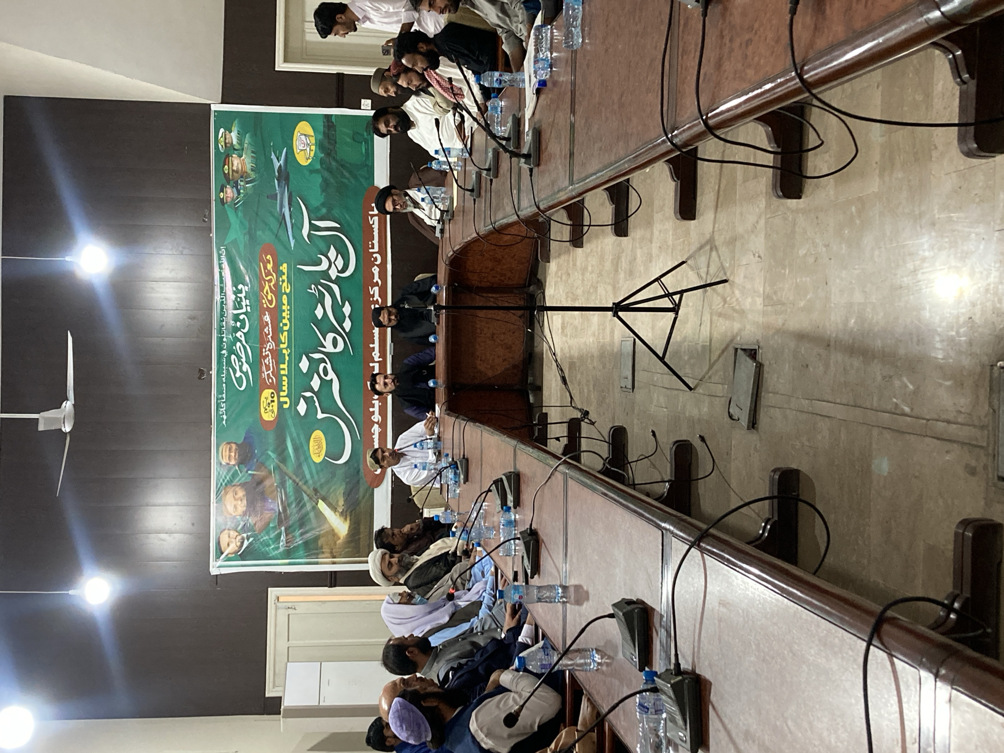 A group of men are seated around a long table in a room with dark wood paneling on the walls and a banner hanging in the background. The banner appears to be promoting or commemorating a specific event or cause, with images of military figures and an airplane, along with Arabic text. The men at the table are dressed in a mix of traditional and modern attire, some wearing turbans and robes, while others are in suits or casual clothing. Microphones and water bottles are placed in front of each person, suggesting a formal meeting or discussion. The table is made of wood and has a decorative edge. Cables from the microphones snake across the tabletop and down to the floor, adding to the organized but somewhat cluttered appearance of the scene. The lighting in the room is somewhat dim, with ceiling lights providing illumination. The scene appears to be located in Quetta City Tehsil, Pakistan, according to the banner.