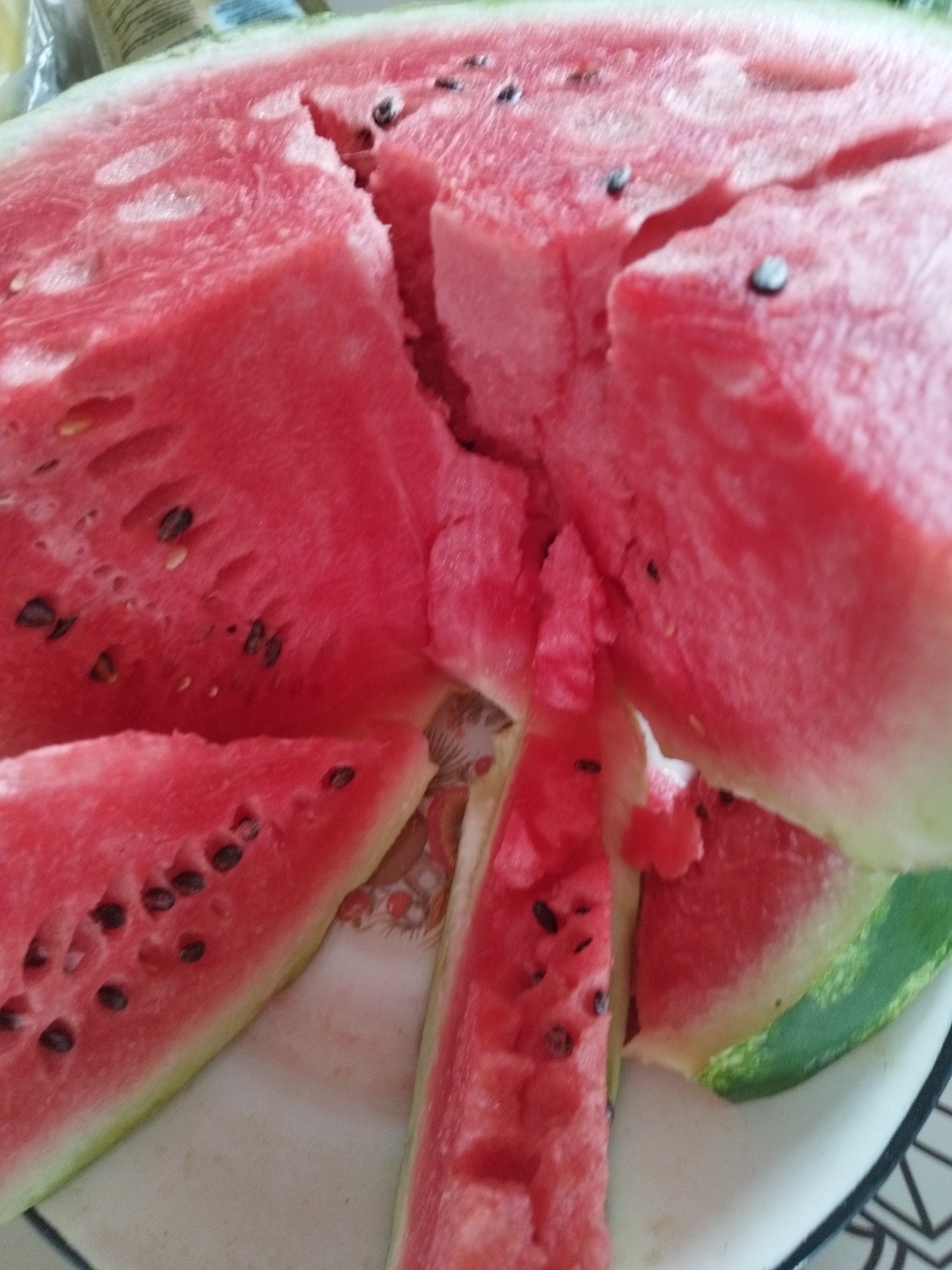 This is a close-up, high-angle shot of several sliced watermelons on a white plate. The watermelon flesh is bright pink and contains numerous black seeds. Some pieces are cut into wedges, while others are broken apart. The rind is green and white. The plate is on a surface with a patterned tablecloth. The image does not contain any elements that would suggest a specific location in Oskemen, Kazakhstan.