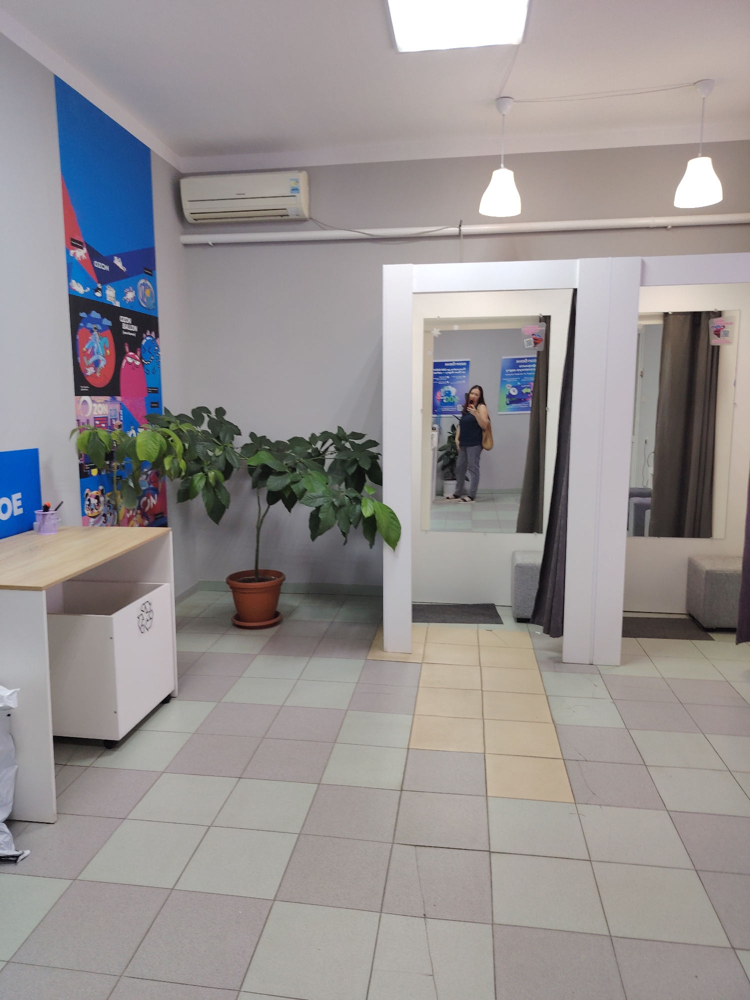 This image depicts a retail fitting room area, likely within a store, with a visible air conditioning unit mounted high on a gray wall. To the left, a blue sign with white text, partially obscured, displays "OE". Adjacent to it is a wooden desk with a white cabinet underneath, containing a trash bin with a recycling symbol and a few pens. A large potted plant with green leaves stands to the right of the desk. The room features a tiled floor with a checkerboard pattern of light gray and off-white tiles. In the background, there is a fitting room area with mirrors and dark gray curtains. A woman is reflected in one of the mirrors, wearing a dark top, gray pants, and carrying a tan bag, holding a phone in her right hand. The scene is well-lit by overhead lighting. The location is identified as Volnoye, Russia.