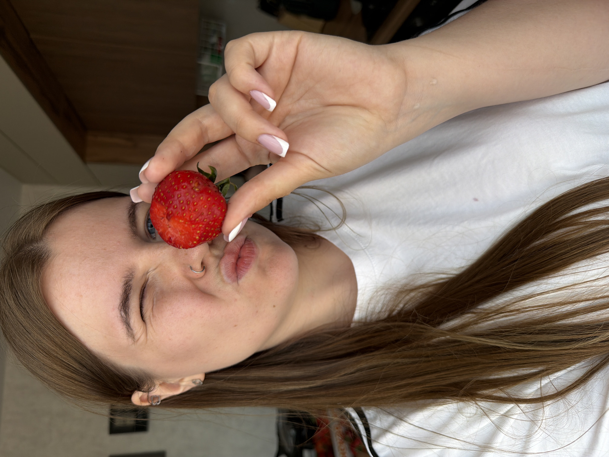 A fair-skinned woman with long brown hair is holding a strawberry in front of her right eye. Her eyes are light blue, and she has a nose ring in her left nostril. She's wearing a white T-shirt. Her nails are manicured with a white French tip. She is pursing her lips. The background is blurry, showing a hint of a wooden wall and some other indiscernible objects. I cannot determine the location in the city of Chaikovsky, Russia, based on the provided information.