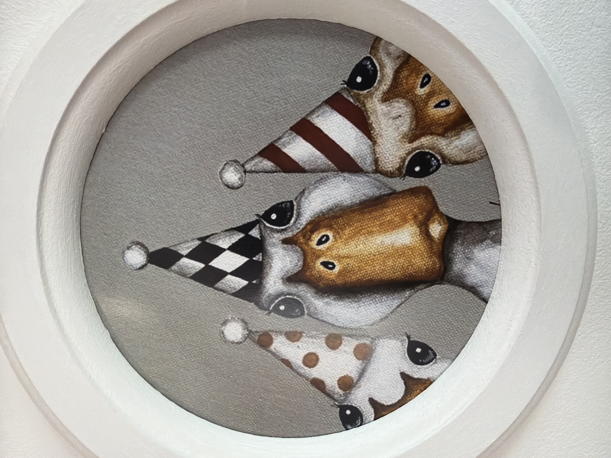 The image shows a close-up of a circular artwork within a white frame. The artwork features three stylized animal faces, likely ducks, wearing party hats. The central duck has a large orange beak and black eyes. The duck to its left wears a polka-dot party hat, and the duck to its right wears a striped party hat. The background of the artwork is a textured gray. The white frame appears to be part of a larger decorative object or architectural element. There is no visible indication of the city of Chaikovsky, Russia in this image.