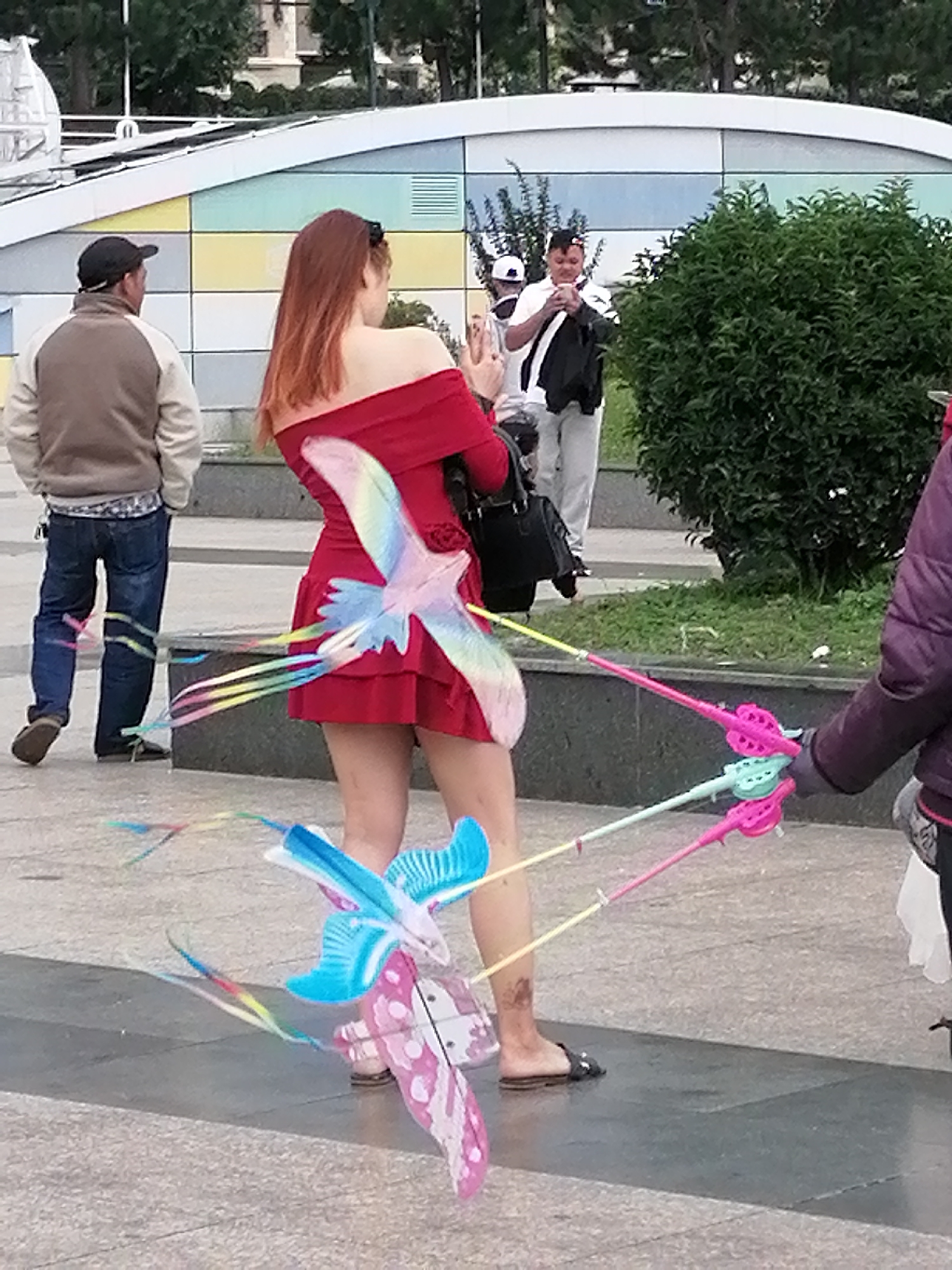 In Da Lat, Vietnam, a woman in a red off-the-shoulder dress is holding two kite reels, each with a kite attached. The kites are shaped like birds, one blue and white, the other a pale pink and white with a patterned tail. Colorful streamers trail from the kites, caught in the breeze. The woman's hair is long and reddish-brown, and she is wearing sandals. Behind her, a man in a light brown jacket and jeans walks away from the camera. Further in the background, another man in a white shirt and black vest is looking down at his phone. The scene appears to be outdoors on a paved area, with a low, colorful wall and lush green bushes in the background. The lighting suggests it is daytime.