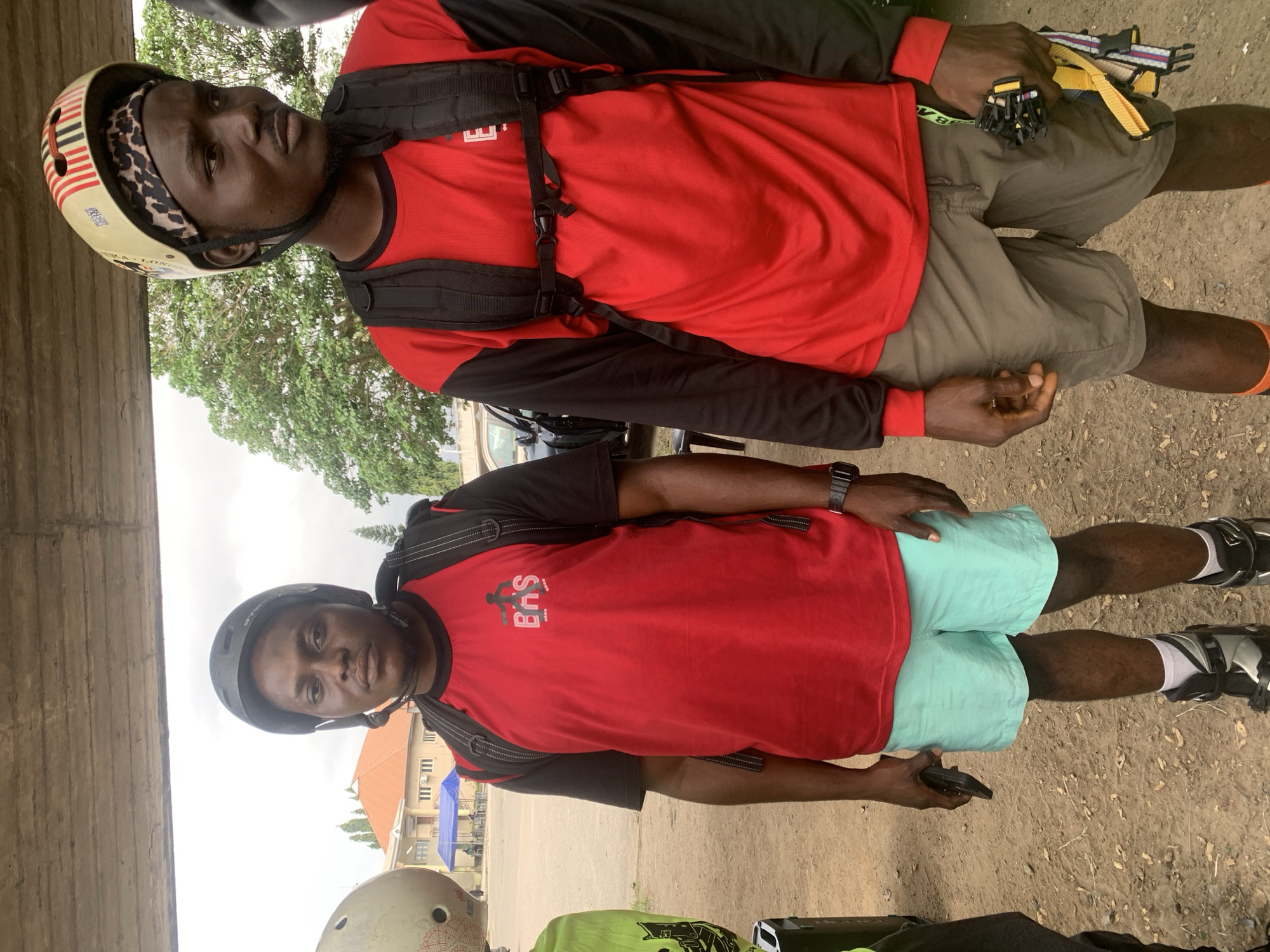 Two men, likely participants in a rollerblading or similar sport, are standing next to each other. The man on the left wears a black helmet, a red t-shirt with black sleeves, and light blue shorts. He is holding a black mobile phone in his left hand and appears to be looking at the camera. The man on the right wears a helmet with red and white stripes and a leopard print bandana. He has a red shirt with black sleeves and grey shorts. He is carrying a black backpack and is holding a bunch of yellow and black straps in his right hand. Both men are wearing protective gear on their legs, suggesting they are prepared for an activity.
The background shows a paved area with some dirt and gravel. There are trees and buildings in the distance, consistent with an urban or semi-urban environment. The sky is overcast. The scene is likely located in Alagbede, Nigeria, given the context. The men are positioned under a concrete structure, possibly a bridge or an overpass.