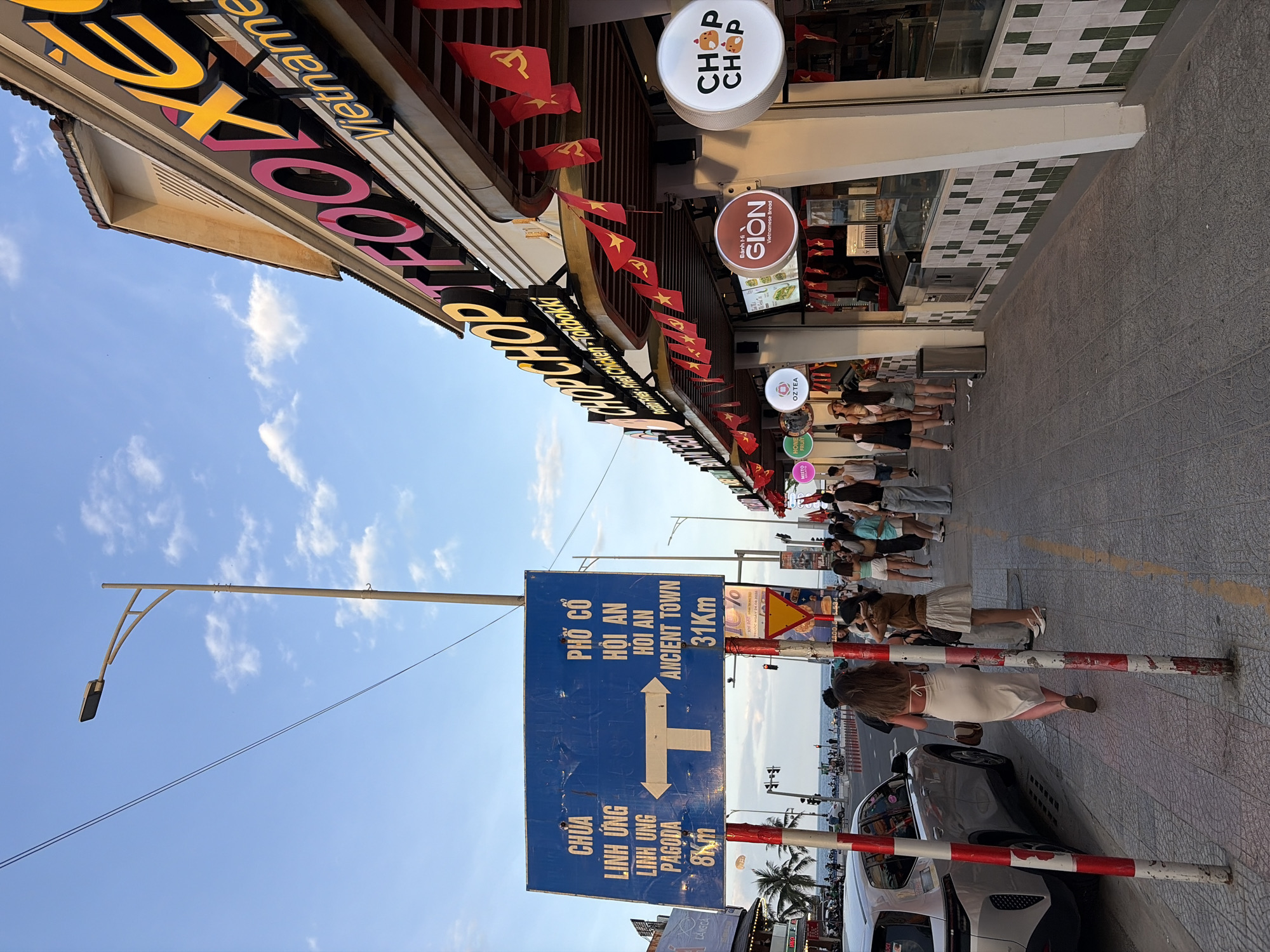 The image depicts a street scene in Da Nang, Vietnam, likely near the beach. A prominent blue directional sign with white text and arrows indicates distances to "Chùa Linh Ứng Pagoda" (8km to the left) and "Phố Cổ Hội An Ancient Town" (31km to the right). The sign is mounted on two poles. To the right, the facade of a building features a large, brightly lit sign that reads "CHOP CHOP THAI FOOD VIETNAM." This establishment is decorated with numerous small red flags with yellow stars, a common symbol in Vietnam. Several people are walking on the sidewalk in front of the building, and a silver SUV is parked on the left side of the street. The sky is blue with scattered clouds, suggesting it is late afternoon or early evening. The overall atmosphere appears to be a lively commercial area.