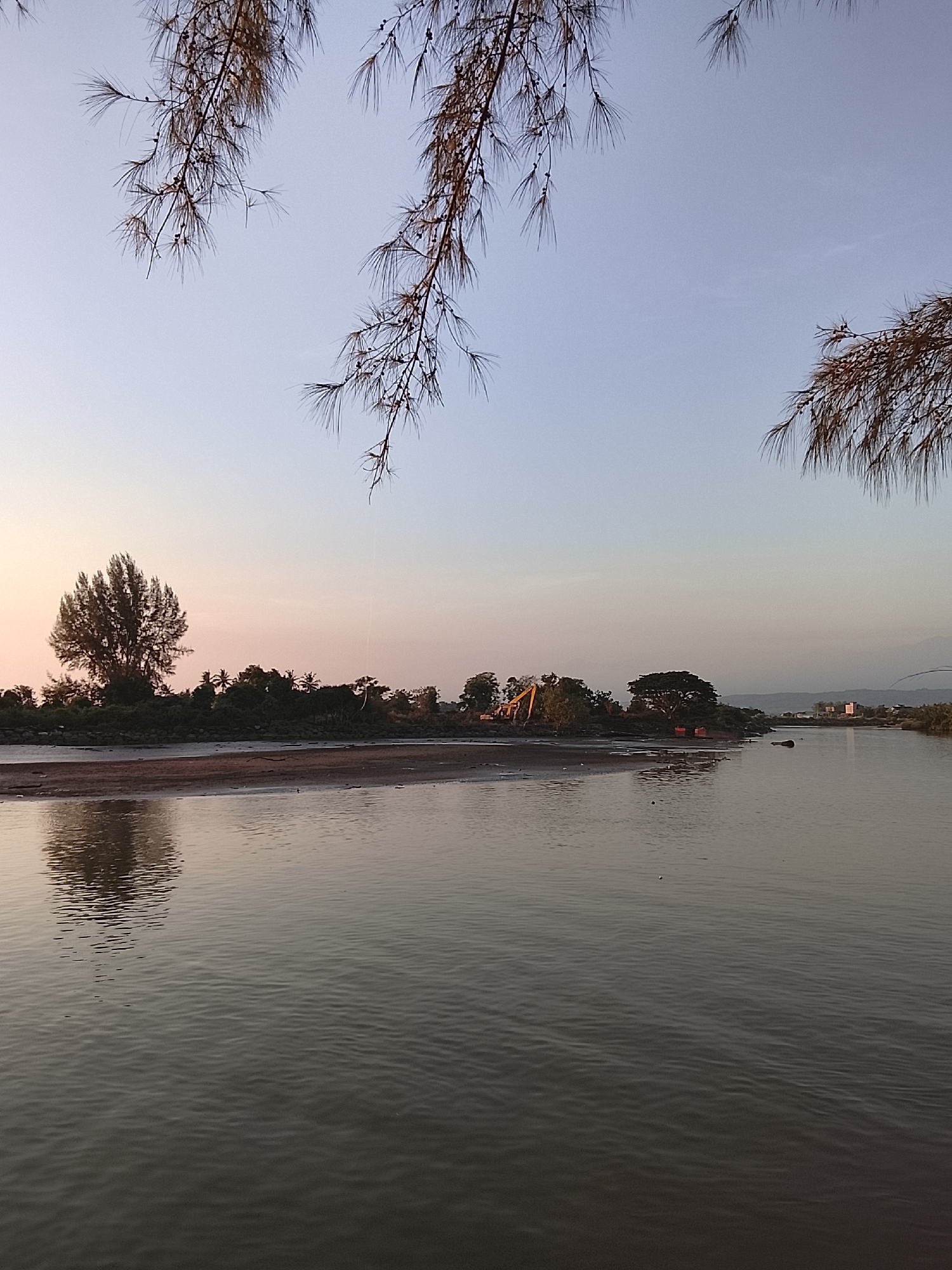 The image depicts a serene river scene in Reudeuep, Indonesia, during what appears to be early morning or late evening due to the soft, pastel colors in the sky. The foreground is dominated by a tranquil river with gentle ripples reflecting the muted light. The riverbanks are lined with lush green trees and vegetation, creating a natural barrier between the water and the land.
On the left side of the riverbank, a prominent, tall tree stands out, its silhouette sharp against the hazy horizon. Further along the bank, the hints of construction activity are visible, with a yellow excavator partially obscured by trees, suggesting ongoing development or maintenance work. A few red structures, possibly boats or small buildings, are also present near the water's edge.
The sky transitions from a pale pink and orange near the horizon to a soft blue as it ascends. The branches of a pine-like tree frame the top of the image, with their needle-like leaves adding a natural element to the composition. The overall atmosphere is one of peacefulness and natural beauty, with a touch of human presence indicated by the construction equipment. There are no people visible in the image.