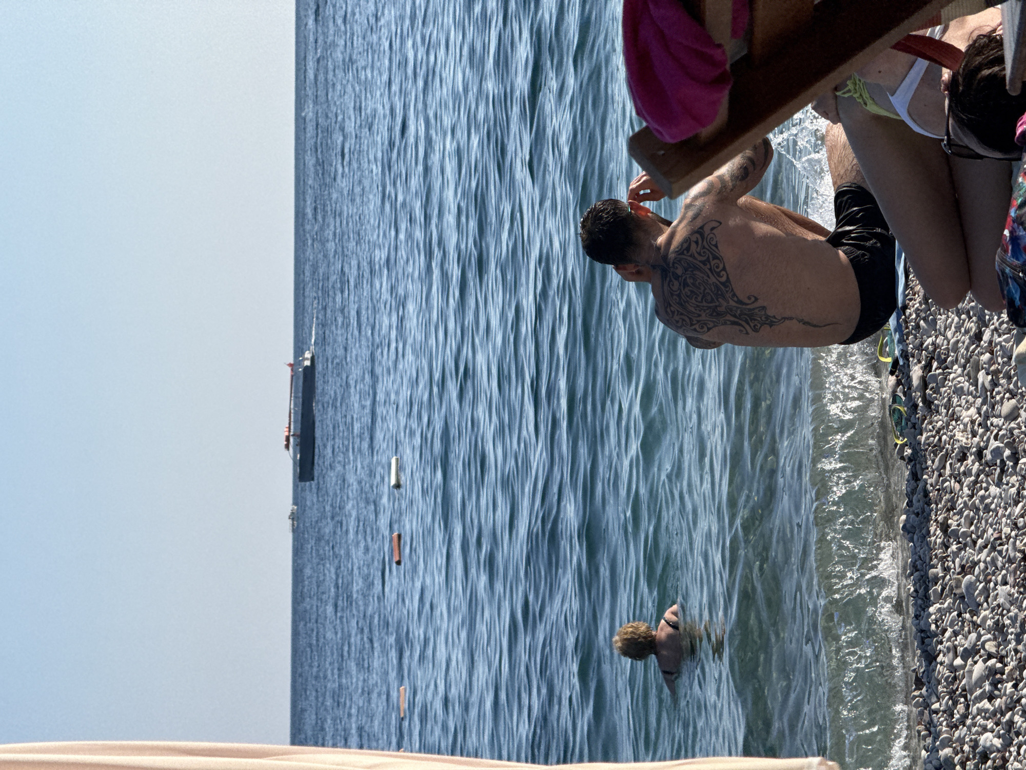 The image shows a group of people enjoying a sunny day at a pebble beach in Budva, Montenegro. The sea is a vibrant blue, with small waves lapping at the shore. A man with a large, intricate tattoo covering his back is partially submerged in the water, looking out towards the sea. Nearby, a person with curly hair is swimming in the water. In the background, a small boat can be seen in the distance. The beach itself is covered in smooth, grey pebbles, and there is a wooden structure in the foreground, possibly a pier or boardwalk. The overall atmosphere is one of relaxation and enjoyment of a summer day. The sky is clear and blue, suggesting pleasant weather. The sunlight is bright, casting reflections on the water and highlighting the textures of the pebbles and the water's surface. The scene appears to be captured in the late morning or early afternoon, given the intensity of the sunlight.