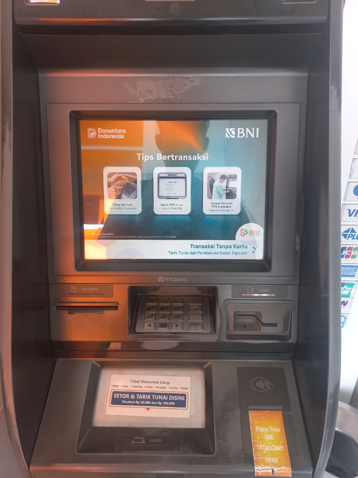 This is a medium shot, eye-level, portrait-oriented photograph of a BNI ATM machine in Baturetno, Indonesia. The ATM has a screen displaying "Danantara Indonesia" and "BNI" logos, along with tips for transactions such as "Tutup jari saat melakukan transaksi" (Cover your fingers when making a transaction) and "Ganti PIN Anda secara berkala" (Change your PIN regularly). It also shows a prompt for "Transaksi Tanpa Kartu" (Cardless Transaction). Below the screen, there is a receipt slot and a keypad with numeric buttons and function keys like "CLEAR" and "ENTER". Further down, there is a slot for card insertion and a "CARD" label. On the right side of the ATM, there is a contactless payment area with a "TapCash" sticker. A sticker in Indonesian that says "SETOR & TARIK TUNAI DISINI" (Deposit & Withdraw Cash Here) is visible, indicating that the machine accepts cash deposits and withdrawals in denominations of Rp 50,000 and Rp 100,000. The overall environment appears to be an indoor or semi-outdoor setting, given the lighting conditions. There are no people visible in the immediate vicinity of the ATM. The ATM is a typical gray, metallic machine with some signage and labels indicating its functions and brand.