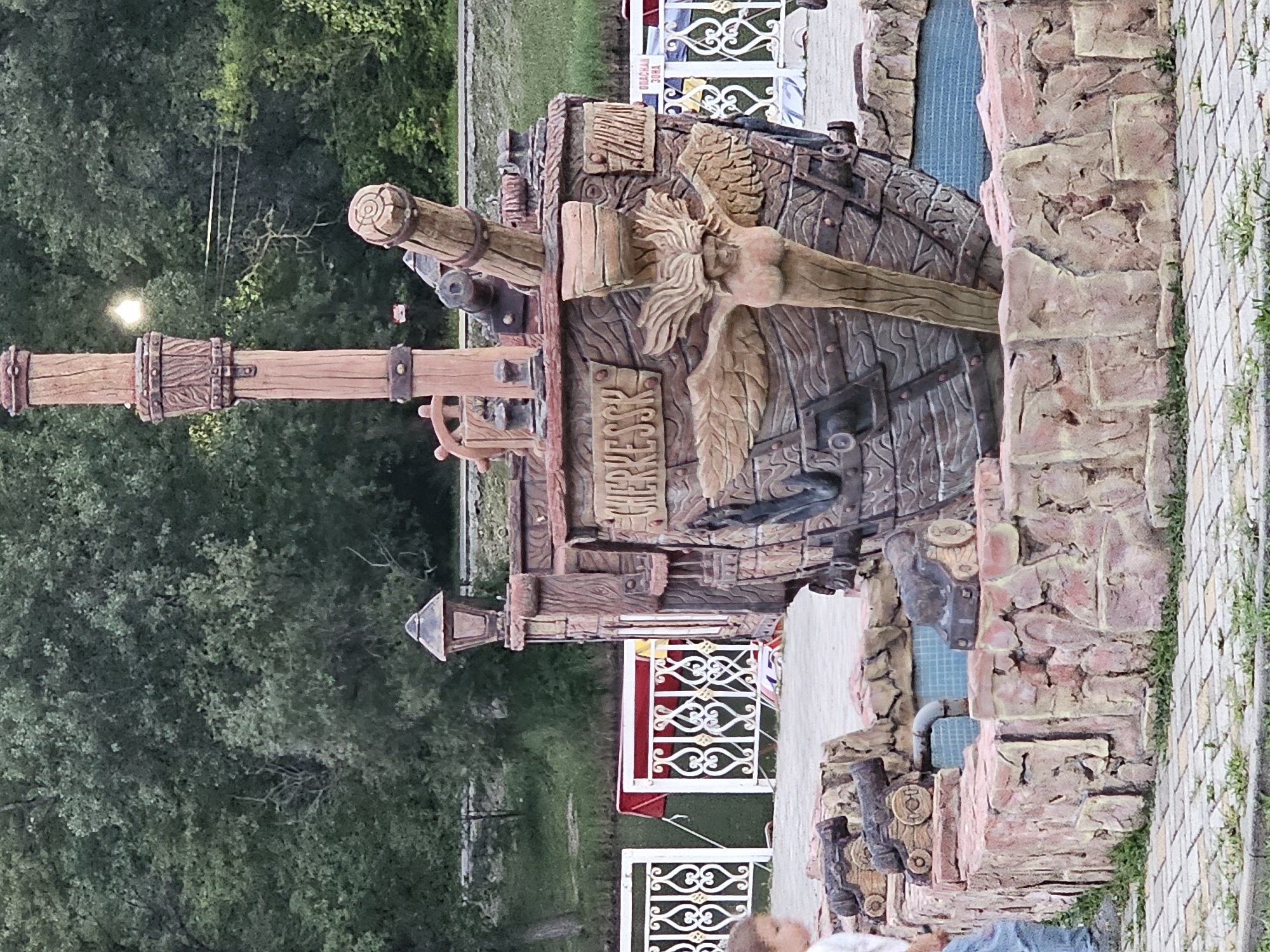 In Cherkessk, Russia, a decorative fountain resembling a pirate ship is situated outdoors. The ship's hull, crafted from rough-hewn wood and stone textures, features a carved siren figure with wings. Two cannons are mounted on the ship's deck, and a steering wheel is visible. The ship sits within a rock-like structure that forms the fountain's basin, with water visible within. In the foreground, a young child with light brown hair, wearing a white shirt and blue jeans, is partially visible on the left, facing away from the camera. The background consists of dense green trees and a body of water, possibly a lake or river. White ornate fencing separates the fountain area from a walkway or park.