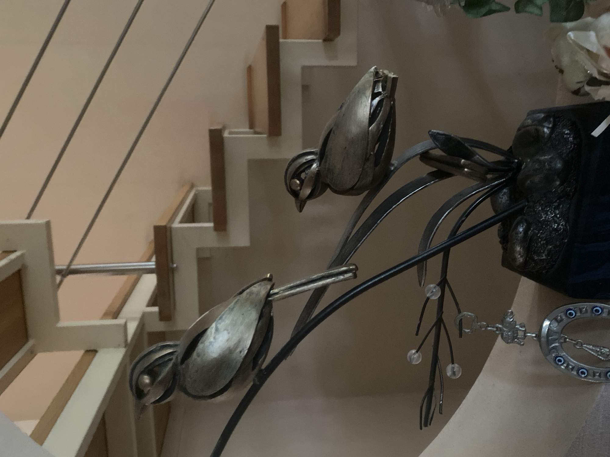 The image shows a decorative metal sculpture of two birds perched on a branch. The sculpture is placed in an indoor setting, likely a home, with a staircase visible in the background. The birds are metallic and appear to be made of brushed or polished metal, with detailed feathering and eye features. One bird is larger and positioned on the left, while a smaller bird is on the right. The branch is also made of dark metal and has small, clear, bead-like embellishments that could represent dew drops or berries. A hanging charm with a horseshoe and an elephant figurine is attached to the branch. The overall ambiance suggests a decorative display in a residential interior. There is no indication of the city of Kharkiv, Ukraine, in the image.