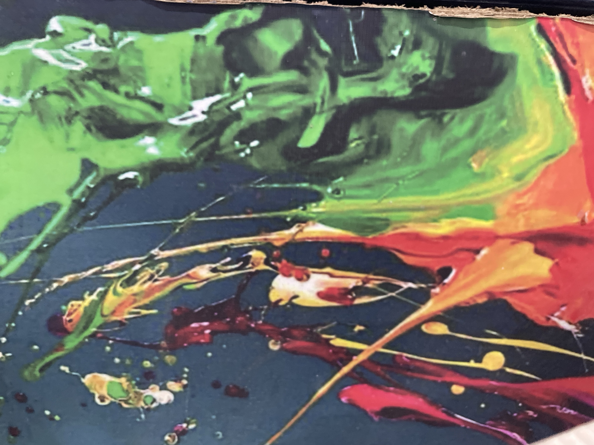 The image shows a close-up of an abstract painting with vibrant colors. Prominent colors include green, black, red, orange, and yellow. The paint appears to be applied in drips, splatters, and streaks, creating a chaotic and dynamic visual effect. A dark blue or black background is visible between the colored elements. There are no recognizable people or objects depicted in this image. There is no indication of any location in the city of Karshi, Nigeria.