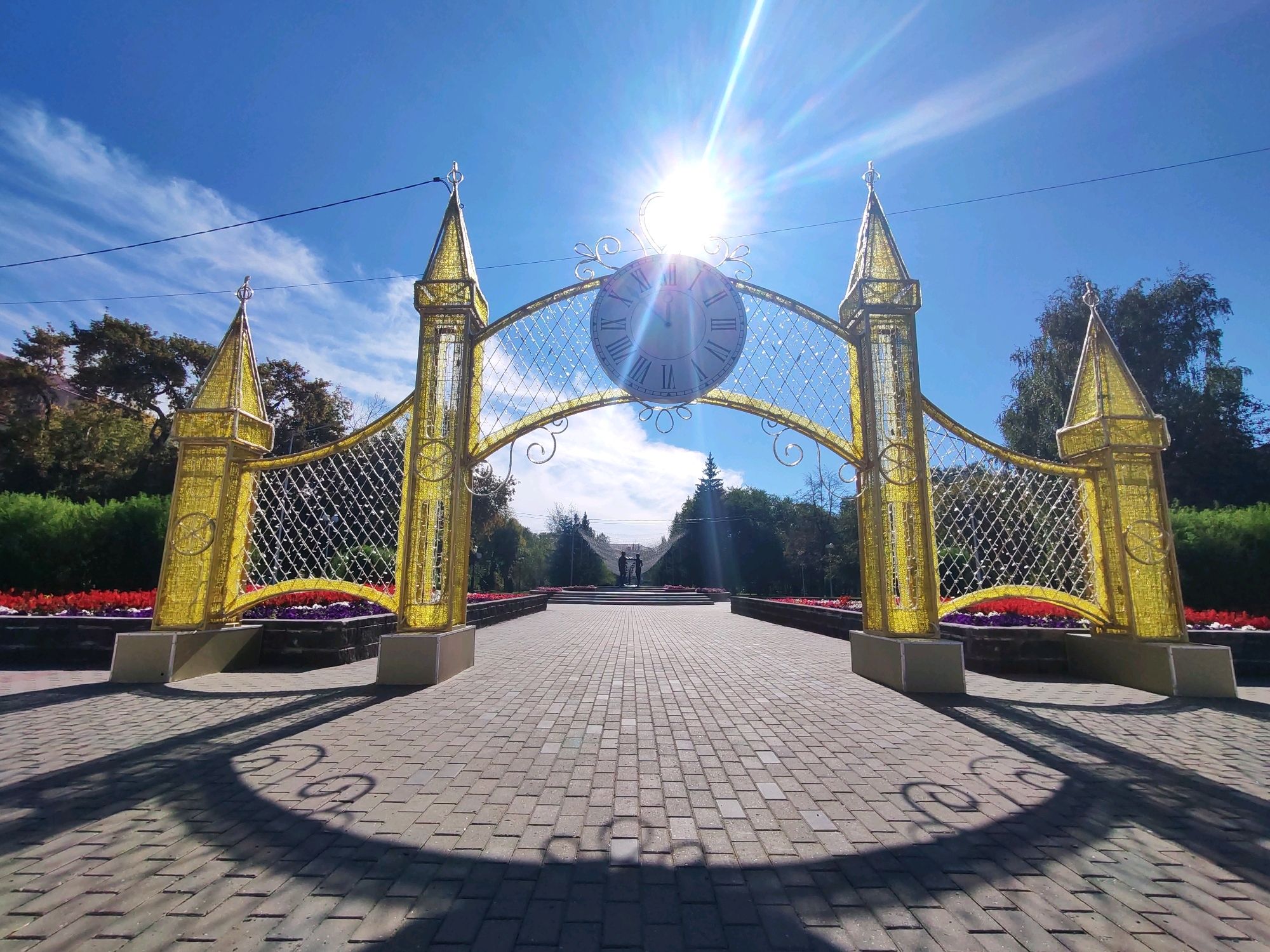 An eye-level, wide-angle shot captures a decorative golden archway with a clock face and pointed turrets, situated in a park. The archway is adorned with a patterned grid and lights, and its base is supported by beige rectangular structures. The ground in front of the archway is paved with light gray bricks, casting distinct shadows from the archway's ornate design onto the surface. Beyond the archway, a pathway leads to a set of stairs with a monument at the top, where two figures stand facing each other. The background is filled with lush green trees and bushes, with a vibrant flowerbed of red and purple flowers flanking the pathway on either side. The sky above is a clear, bright blue with wispy white clouds. The sun is positioned high in the sky, casting a strong glare and sunbeams that extend across the scene.