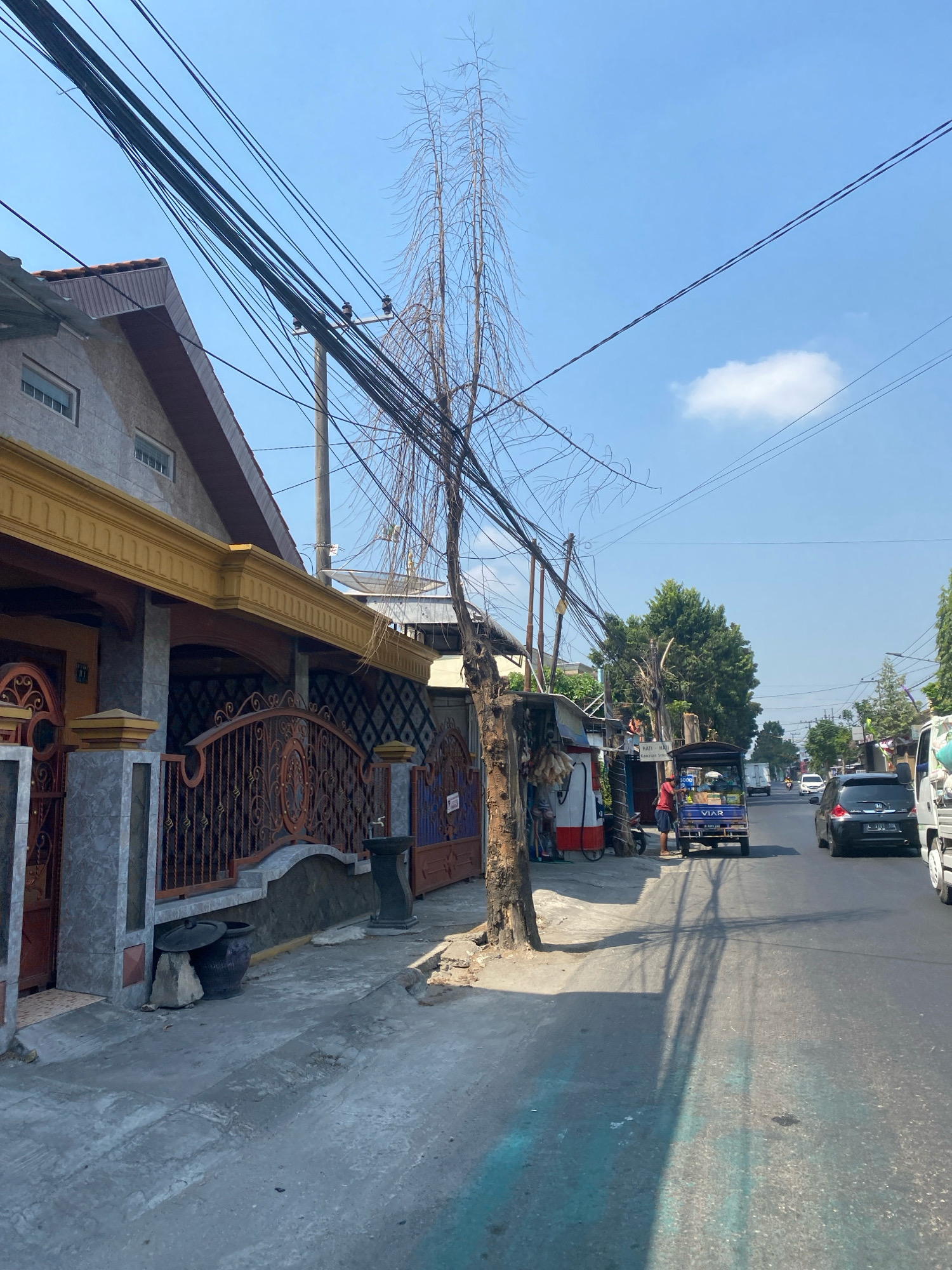 The image captures a street scene in the city of Kediri, Indonesia, on a bright, sunny day. A residential building with a yellow decorative trim and an ornate iron fence lines the left side of the street. In front of the fence are two large dark ceramic pots, one with a lid, and a stone. A tall, dry tree with numerous thin branches stands prominently near the sidewalk, its silhouette stark against the blue sky. Overhead, a complex network of black electrical wires crisscrosses the frame.
The street itself is paved, with vehicles present. A blue and white auto-rickshaw, identified by the "VIAR" logo, is parked on the right side of the street. A person in a red shirt is standing next to it, appearing to interact with the vehicle. Further down the street, other vehicles, including a black car and a white truck, are visible, suggesting ongoing traffic. A small roadside stall with red and white accents is situated on the left, near the dry tree, with goods displayed. Lush green trees are visible further down the street, contrasting with the dry tree in the foreground. The shadows cast by the overhead wires stretch across the asphalt, indicating the strong sunlight. The overall environment suggests a typical urban setting in Indonesia, characterized by residential structures, overhead utility lines, and active street life.