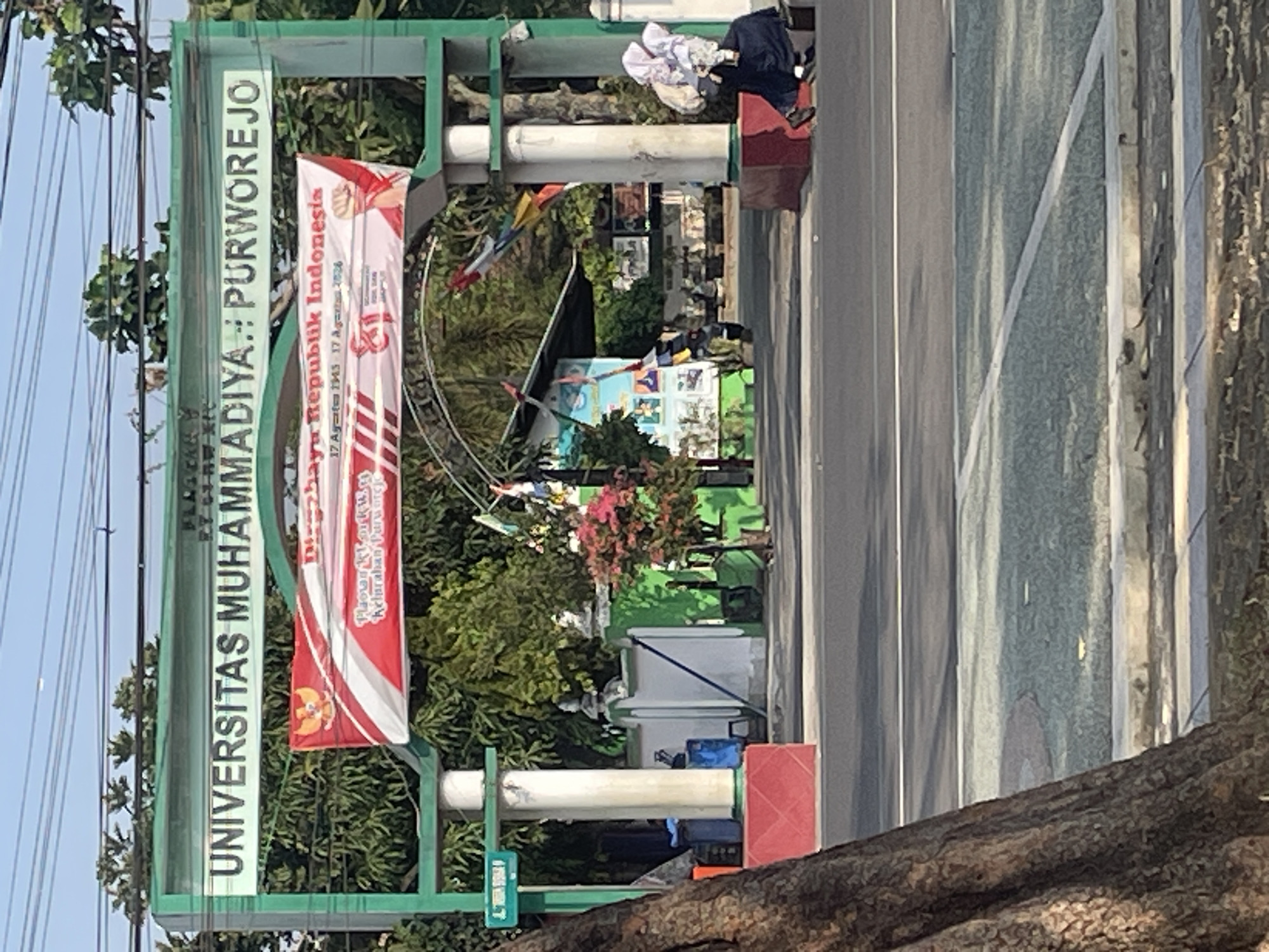 The image shows the entrance gate of Universitas Muhammadiyah Purworejo, located in Purworejo, Indonesia. The gate is a green archway with the university's name, "UNIVERSITAS MUHAMMADIYA PURWOREJO," displayed in white lettering on a light green background. Below the university name, a banner is hung, featuring the Indonesian flag colors (red and white) and text related to the Indonesian Republic's Independence Day. The banner also includes an image of a clenched fist.
In the background, behind the gate, there are trees and buildings associated with the university. A white wall with green accents is visible, along with a small tree with pink flowers. There are several people present. On the right side of the image, two individuals are sitting on a red-lined platform near the gate, one of whom is wearing a white hijab and dark clothing. Further inside the university grounds, other people can be seen walking.
In the foreground, a large tree trunk is prominent on the left side. The scene appears to be a street or road in front of the university entrance, with a paved surface and what looks like a pedestrian walkway or grass area on the right. The sky is clear and blue, suggesting it is daytime.