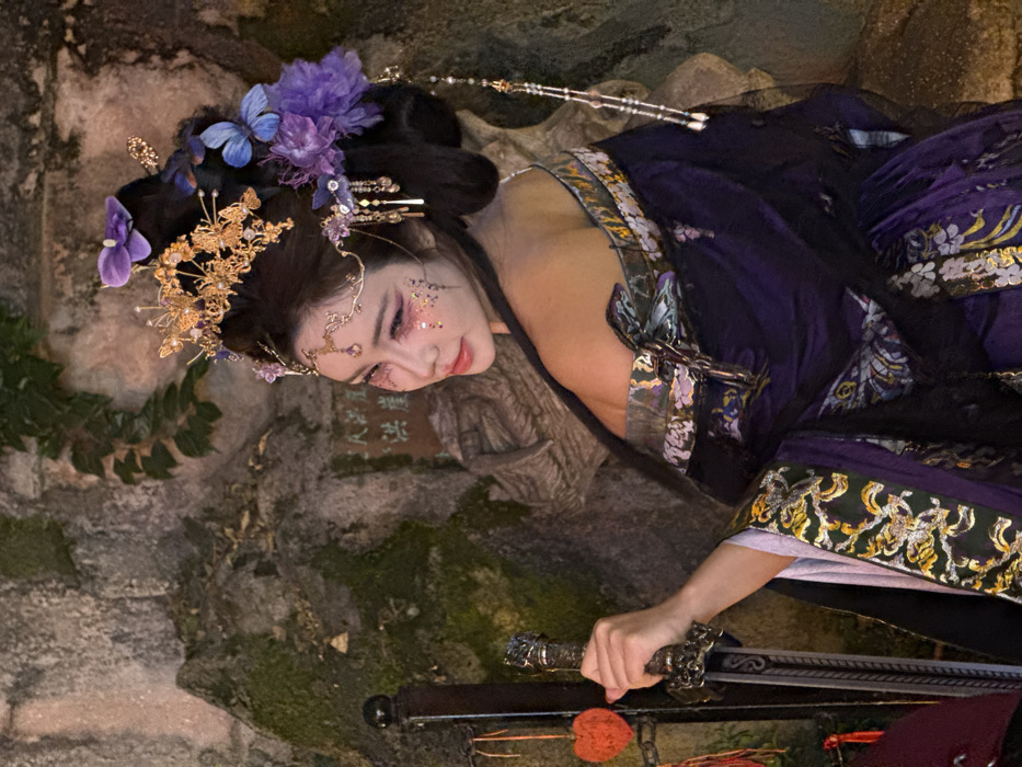 A person in traditional attire poses with a sword against a stone background. The individual, adorned with floral headpieces and glittery makeup, wears a dark purple and gold garment with ornate details. The sword, held in their right hand, rests in a scabbard. The surrounding environment includes mossy stone structures, illuminated by soft lighting, which creates a fantastical atmosphere.