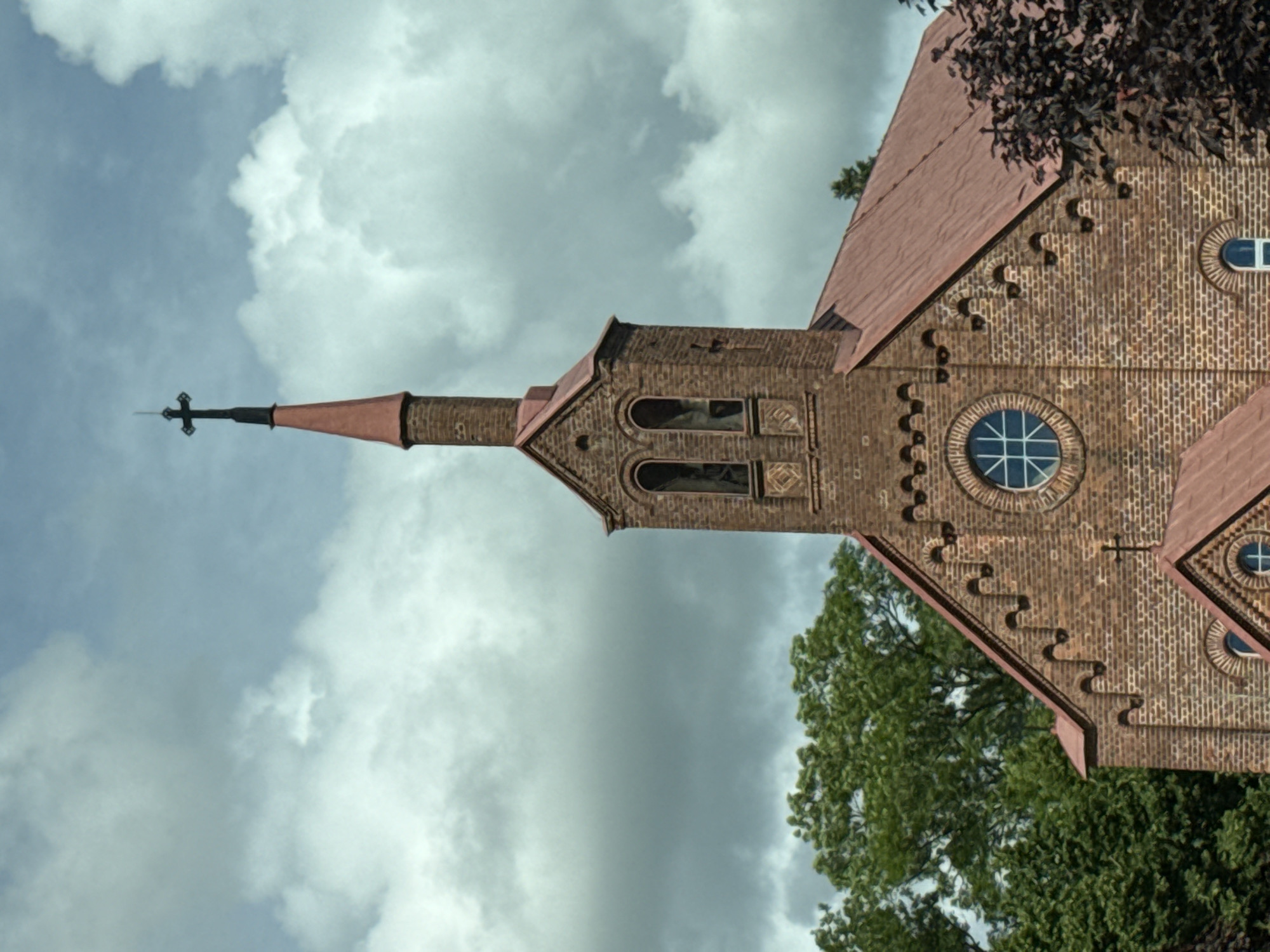 The image shows the exterior of a brick church with a red, sloped roof. A tall, slender spire with a cross on top rises from the church. The spire is made of brick and topped with a conical, copper-colored roof. Below the spire, the church has a bell tower with two arched openings. A circular window with a cross-shaped mullion is visible on the main body of the church. The church is surrounded by green trees and a cloudy sky. This is the Holy Trinity Catholic Church in Šilutė, Lithuania.