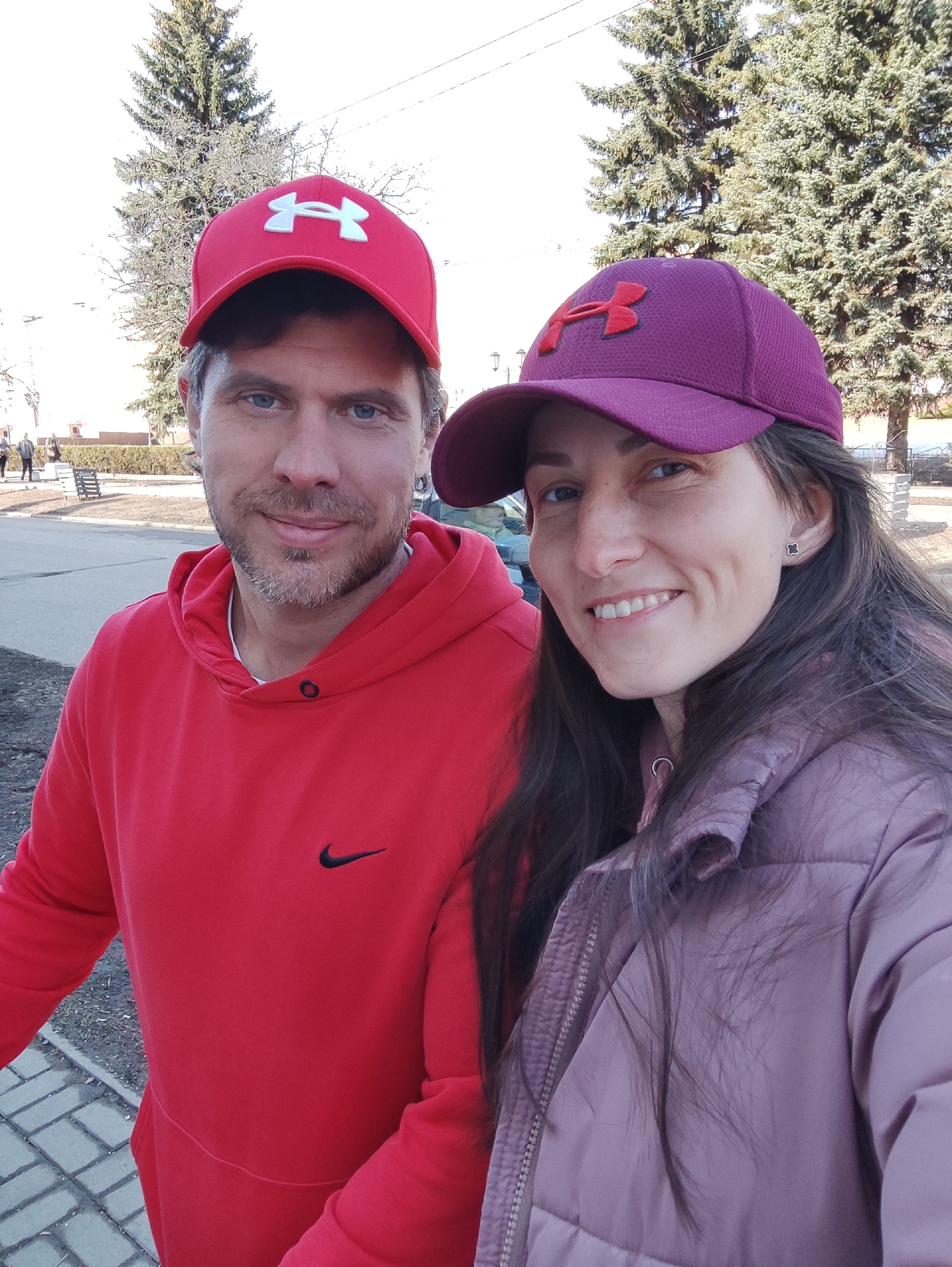 A man and a woman are posing for a selfie outdoors. The man is on the left, wearing a red hooded sweatshirt with a black Nike logo and a red baseball cap with a white Under Armour logo. He has short brown hair, blue eyes, and a slight beard. The woman is on the right, wearing a mauve-colored puffy jacket and a matching baseball cap with a purple Under Armour logo. She has long brown hair and is smiling.
The background shows a paved area, bare trees, and a few people walking in the distance. The photo was taken in Izhevsk, Russia.