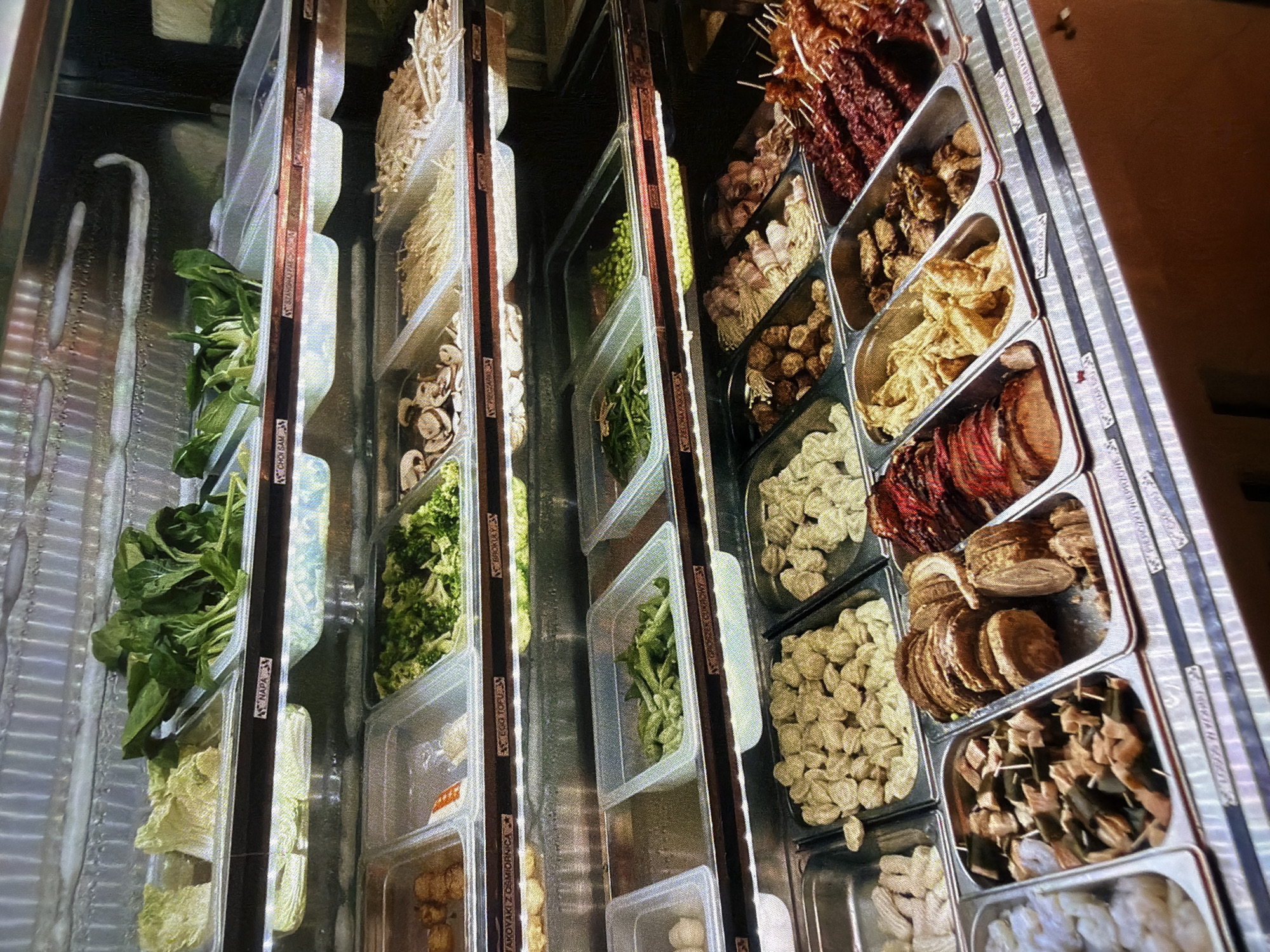 The image shows a close-up, eye-level view of a refrigerated buffet display case filled with various food items. The display case has multiple shelves, each containing stainless steel pans filled with different ingredients. The items appear to be arranged for a self-serve meal, possibly hot pot or a similar cuisine.
On the top shelf, to the left, are chopped napa cabbage and what appears to be bok choy. To the right of the bok choy are more green leafy vegetables, possibly spinach.
The second shelf from the top contains sliced mushrooms, a tray of enoki mushrooms, and another tray of thinly sliced, light-colored ingredients that could be bamboo shoots or another type of mushroom.
The third shelf from the top has a variety of items. On the left are what look like small, fried dough balls or dumplings. Next to them is a tray of broccoli florets. Further to the right are sliced shiitake mushrooms. There are also trays of what appear to be bean sprouts and possibly peas.
The bottom shelf of the display case showcases pre-prepared skewers and meat items. On the far left, there are shrimp. Moving to the right, there are skewers with chunks of meat and dark green vegetables, followed by sliced cooked meats that appear to be roast beef or pork. Then there are trays of what look like gyoza or dumplings, and some grilled meat skewers.
The overall environment is clean and well-lit, consistent with a food service establishment. The background is metallic and reflective. There are no people visible in the image, and no discernible location beyond the buffet itself. The quality of the image is good, with clear detail of the food items.