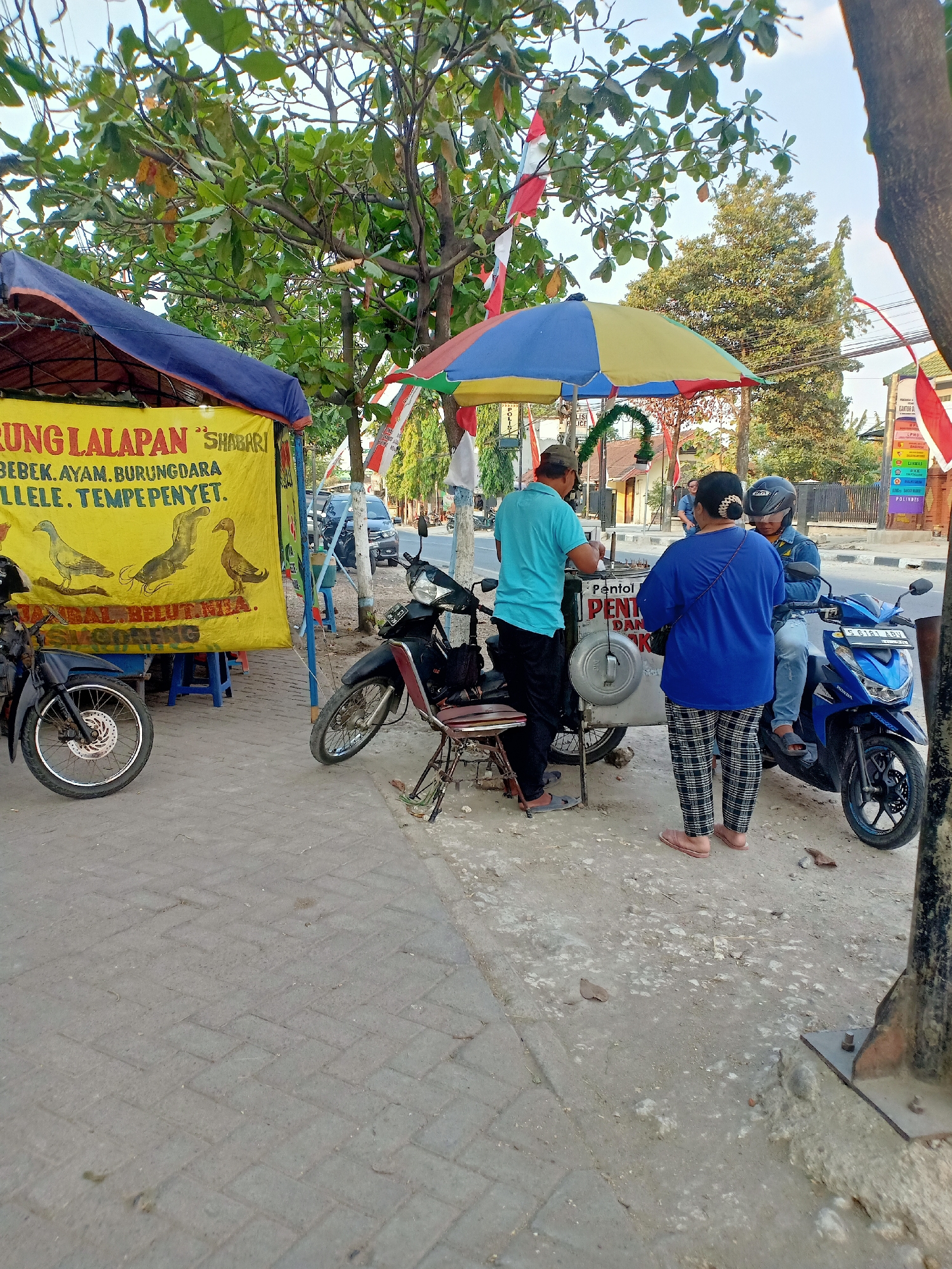 The image depicts a street scene in Sumber Tlaseh, Indonesia. In the foreground, a food cart vendor is serving customers. The vendor, wearing a blue shirt and a cap, is interacting with a woman in a blue shirt and checkered pants. A person wearing a helmet is sitting on a blue motorcycle, facing the cart. The food cart itself is small and white, with a colorful umbrella over it. To the left of the cart, there is a larger yellow tarp tent with various food items advertised on it. Two motorcycles are parked near the food cart and tent. The ground is paved with interlocking blocks. In the background, there are trees, buildings, and a road with passing vehicles. Indonesian flags are visible, suggesting a possible celebration or event. The overall scene is that of a busy roadside eatery.