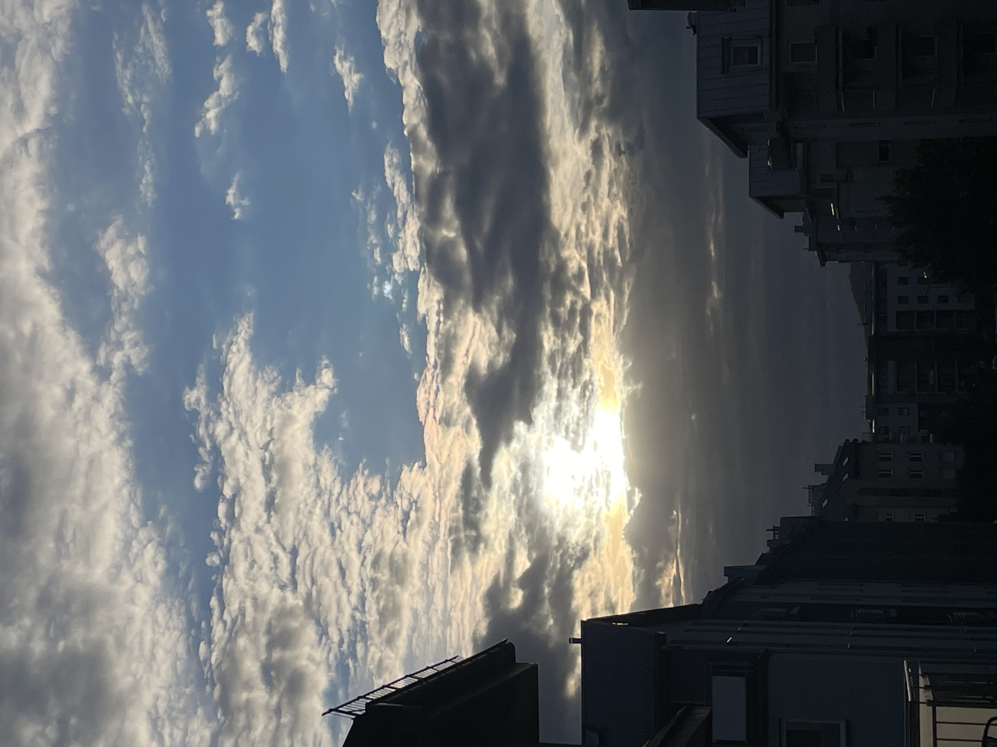 This is a vertical, medium shot of a city skyline under a dramatic sky with the sun breaking through clouds. The sky is a mix of bright blue and gray clouds. The sun is positioned in the center of the frame, partially obscured by dark, heavy clouds, casting a bright glow and illuminating some of the surrounding cloud formations.
In the foreground and midground, silhouetted buildings of various heights form the city skyline. The buildings are dark and appear to be residential or commercial structures with visible windows and rooflines. The perspective suggests the viewer is looking out from a higher vantage point, possibly a balcony or window. The overall lighting is dim due to the clouds, with a strong emphasis on the bright sun. There are no people visible in the image. The environment is urban.