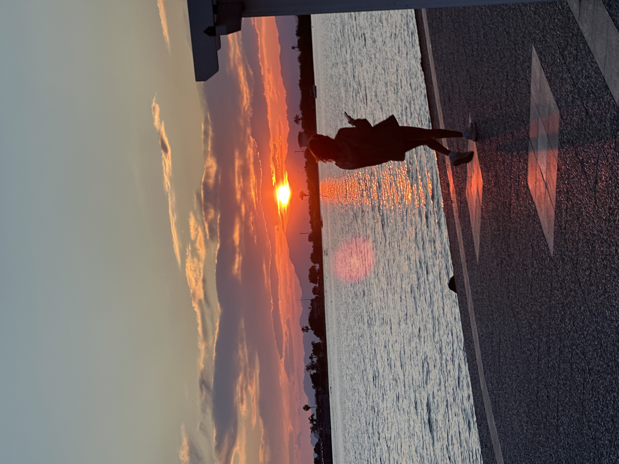 A sunset view over the water from Fethiye, Turkey, shows a person silhouetted, standing on a stone promenade. The sun is setting over a distant tree-lined shore, casting an orange and red glow across the sky and reflecting in the water. The promenade is marked with geometric designs, catching the light.