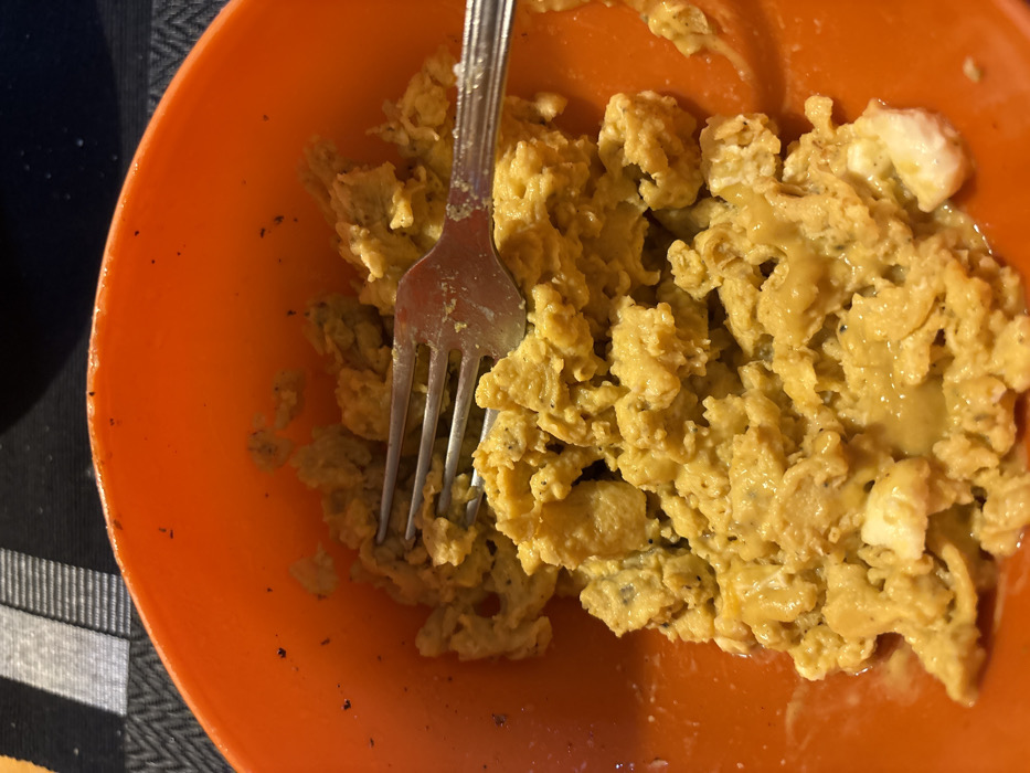 The scene depicts a close-up, elevated view of an orange plastic bowl containing scrambled eggs, situated on a dark surface with a horizontal black and gray striped pattern. The location is the city of Stefan Karadja, Bulgaria.
The scrambled eggs occupy the majority of the bowl, presenting a lumpy, irregular texture with a predominant yellow-orange hue. Visible within the eggs are occasional lighter, off-white segments and numerous dark specks, indicative of seasoning such as ground pepper. A silver-colored metal fork is partially embedded in the eggs, positioned with its tines facing towards the upper left portion of the frame. The fork's handle extends towards the right, resting across the surface of the eggs. The interior rim of the orange bowl exhibits some dark residue or specks similar to those in the eggs. The background behind the bowl is out of focus, showing the aforementioned striped pattern. No individuals are visible.