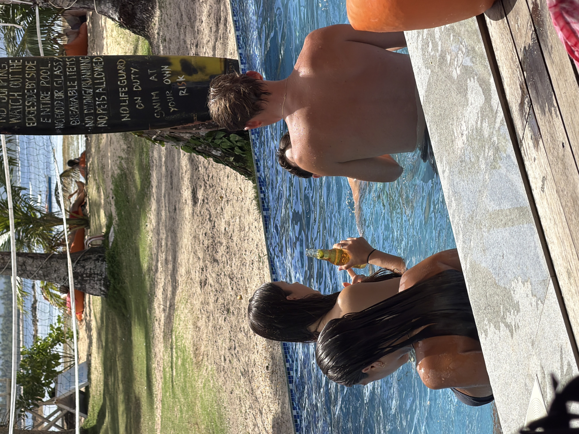 A woman holds a beer bottle in a pool, a man stands behind her, and a sign in the background reads "No Lifeguard on Duty, Swim at Your Own Risk". The scene is likely in General Luna, Philippines, given the location of the sign and pool. The pool is surrounded by sand and palm trees.