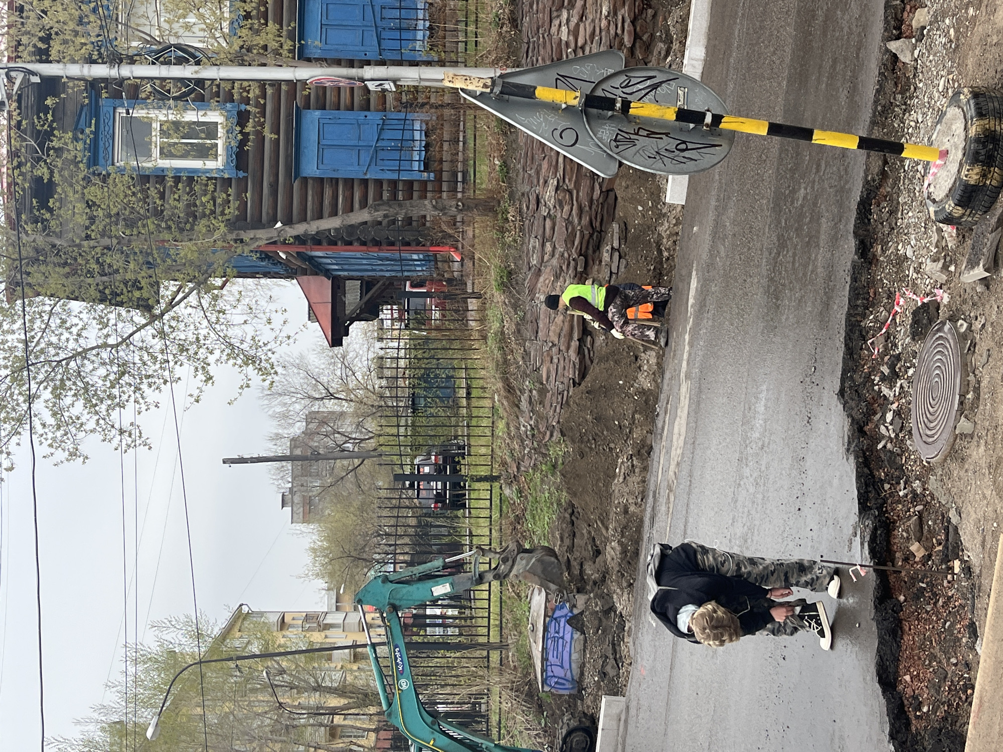The image shows a street repair scene in an urban environment. Two construction workers are working on the street. One is wearing a black beanie and a high-visibility lime green vest. The other is wearing a black jacket, camouflage pants, and black and white shoes. An excavator sits on the side of the road. A street sign is positioned near the edge of the excavation area. A wooden building with blue window frames is visible in the background. A black wrought iron fence is also visible. The location appears to be the city of Unknown, Unknown.