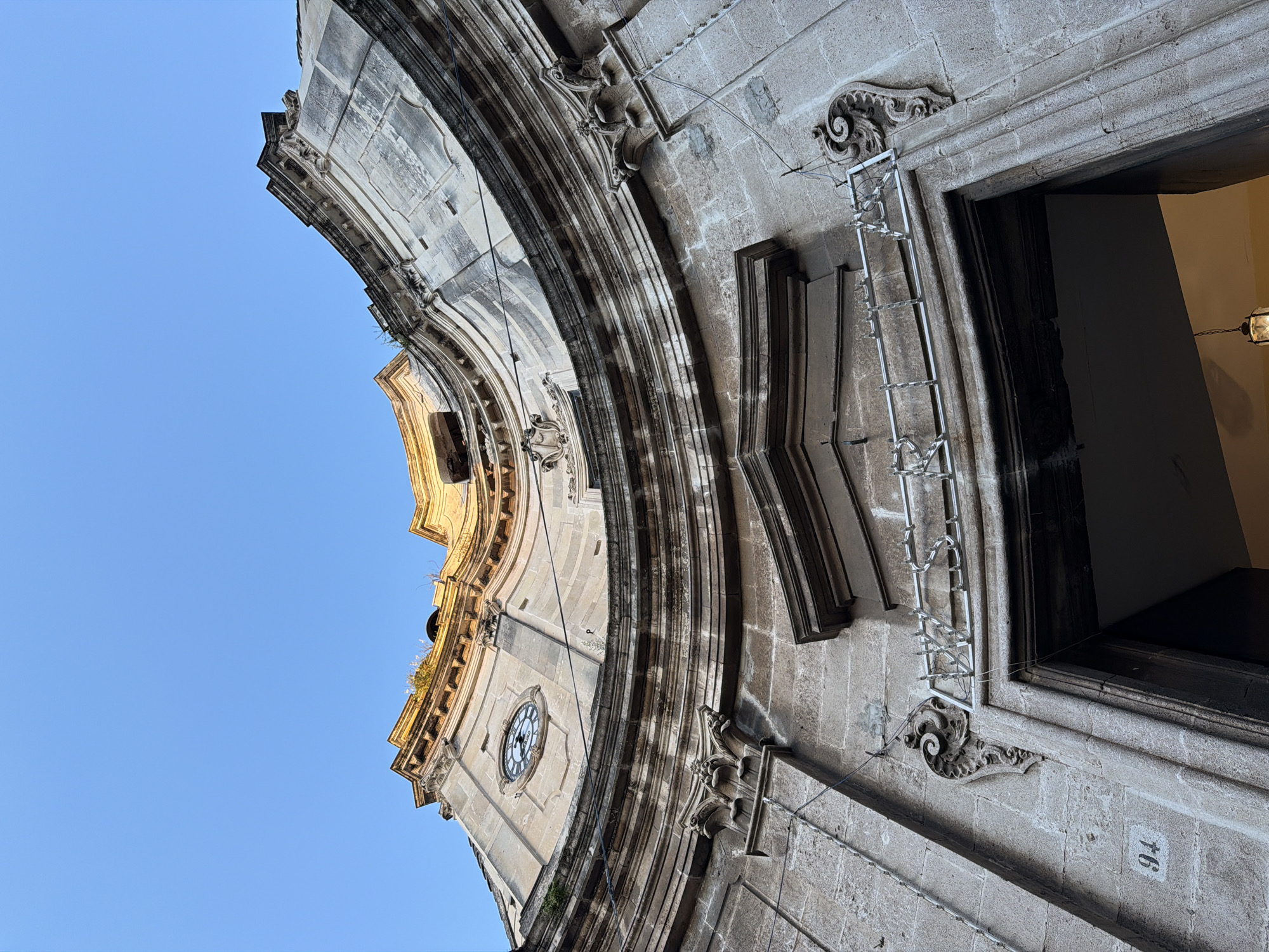 The image shows a section of a building in Acireale, Italy, shot from a low angle, looking upwards. The building is constructed from stone and features ornate architectural details, including curved moldings, carved embellishments, and a clock on the side of the building. The façade also includes a rectangular window or doorway, which is partially visible. A silver ladder is affixed to the side of the building near the window. The number "94" is written in white on the building's exterior. The sky is visible behind the building and is a clear, pale blue color.