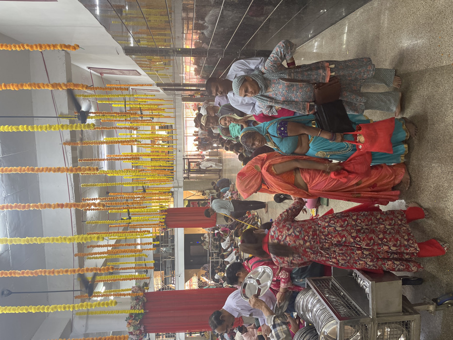 The image shows a large gathering of people in a large, open space. The location is a temple in Panchkula, India, based on the coordinates provided. The ceiling is adorned with strings of yellow marigold flowers. The space is decorated with red drapes. On the right side of the image, people are standing in line. A group of people is sitting on the floor in the left foreground. A group of women are standing in the center foreground. Many of the women are wearing colorful saris. One woman is wearing a red and floral dress. In the left foreground, a man in a white shirt and a woman in a red and floral dress are distributing plates from a cart. Other people are holding plates and waiting to be served.
