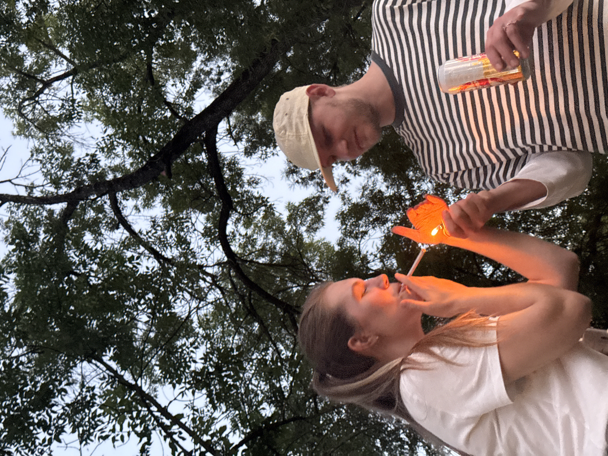 A low-angle shot shows a woman in a white shirt with a ponytail lighting a cigarette from a lighter held by a man in a striped shirt and beige cap. The man is also holding a glass or can. They appear to be outdoors, under trees with visible branches and leaves. The location is not discernible from the image.