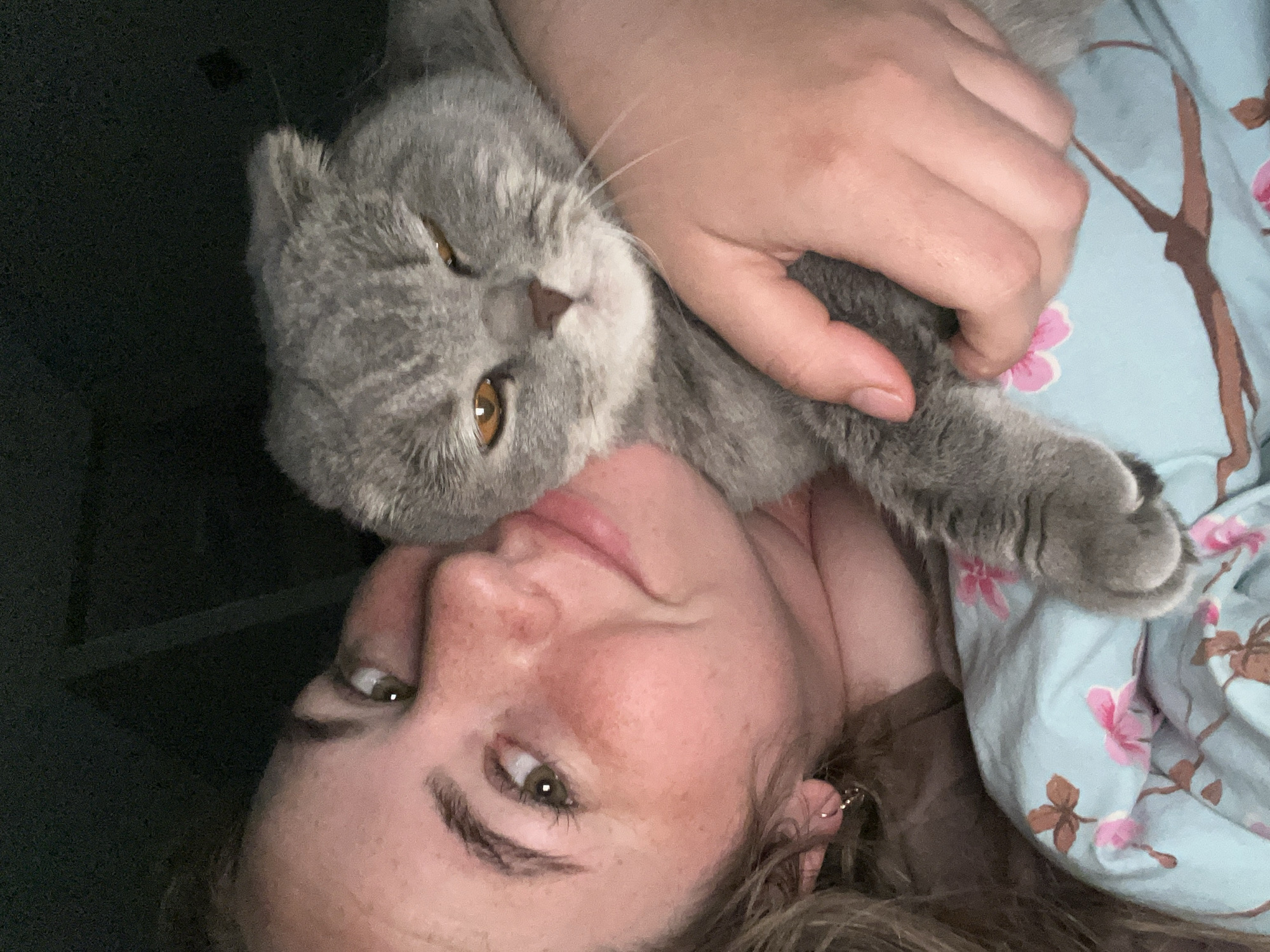 A woman is holding a gray cat. They are lying down on a bed with a blue floral patterned blanket. The woman's face is close to the camera, she is looking at the camera with a slight smile. The cat is nestled against her neck and shoulder. The image is taken indoors. The location in the city of Chaikovsky, Russia is not discernible from the image.