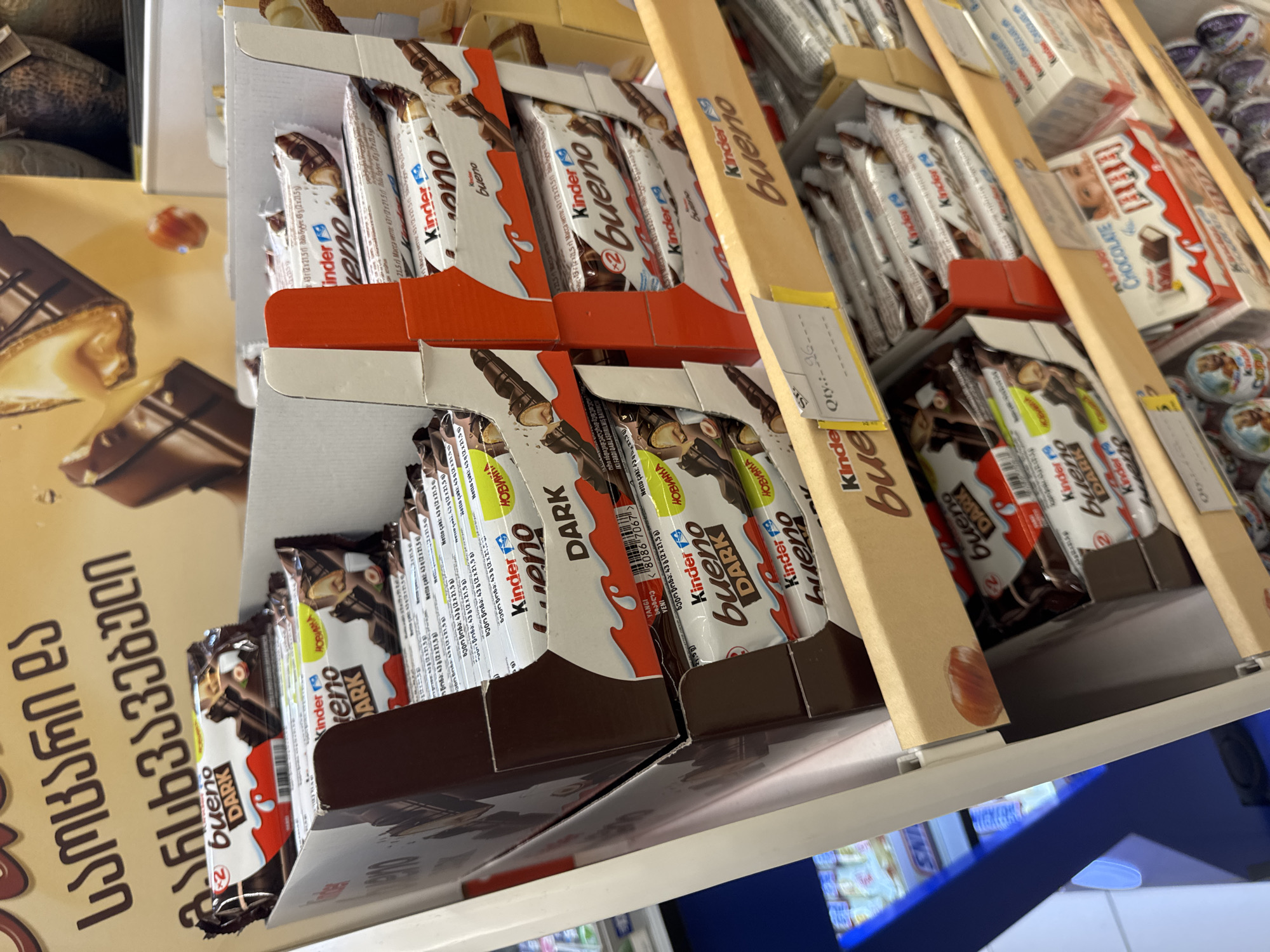 The image shows a retail display of Kinder Bueno chocolates in a store. The products are primarily stacked in cardboard boxes, with some individual wrappers visible. The store appears to be a supermarket or convenience store, with other products and shelving visible in the background, including what looks like a display of Snickers bars on the left and boxes of Kinder Chocolate on the bottom right. The overall lighting suggests an indoor setting. The text visible on the packaging is primarily in Russian and Georgian. The Georgian text at the top of the image can be translated as "Sweets and chocolate" or similar. The location is identified as being in Tbilisi, Georgia.
