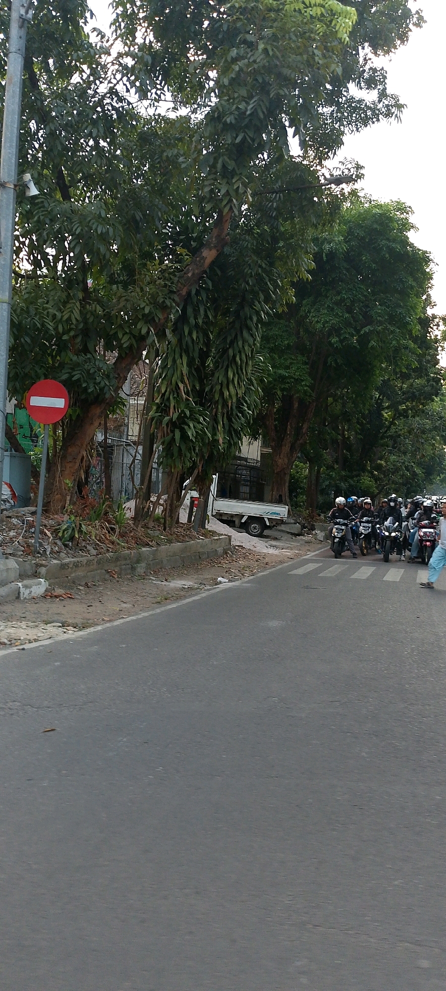 This is a medium shot, taken from a low angle, of a street scene in Bandung City, Indonesia, featuring lush trees lining the road. In the foreground, a gray asphalt road with a white line marking the edge is visible. To the left, a traffic sign with a red circle and a white horizontal bar, indicating "No Entry" or "Do Not Enter," stands next to a concrete utility box. Behind the sign, tall trees with dense green foliage dominate the left side of the frame, with some showing signs of age and epiphytic growth.
In the middle ground, a narrow strip of dirt and rubble separates the road from the trees. A white truck with an open bed is parked on the right side of the road. Further down the road, a procession of motorcycles is seen, with riders wearing helmets and some carrying passengers. The motorcycles are lined up on the right side of the road, suggesting a pause or slow traffic. A person in light-colored pants is walking across a pedestrian crossing on the far right of the image. The background is filled with more trees and hints of buildings, creating a natural and urban blend typical of Bandung. The sky is overcast and bright, suggesting it's daytime.