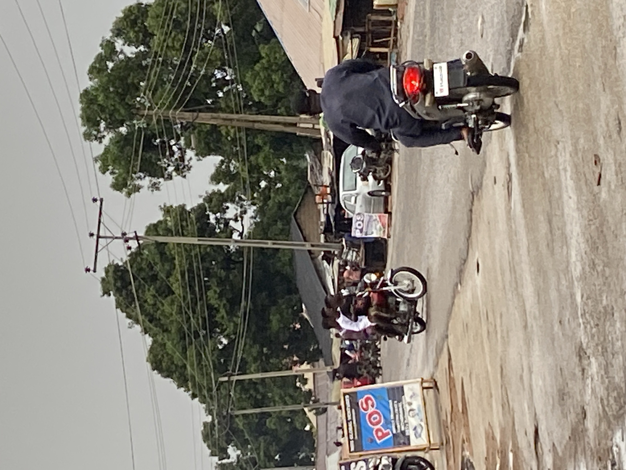 The image shows a street scene in Mararaba, Nigeria. Several people are riding motorcycles along the road. Power lines and poles line the left side of the street, alongside trees and buildings. A POS (Point of Sale) advertisement is visible on the lower right. The street appears to be paved, but with visible wear and tear. The sky is overcast.
